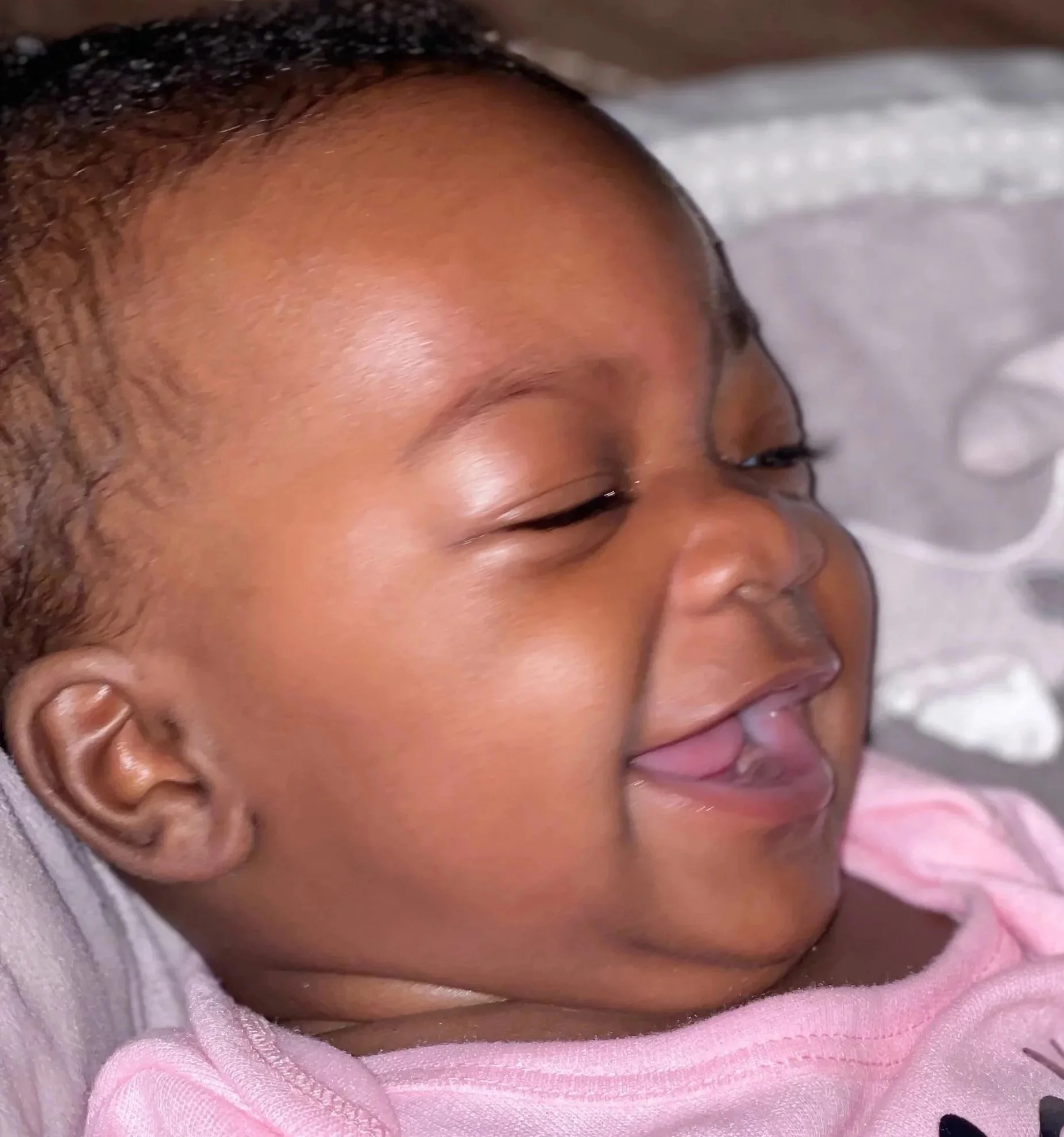 Close-up of a smiling baby with closed eyes, wearing a pink outfit, lying on a white blanket.