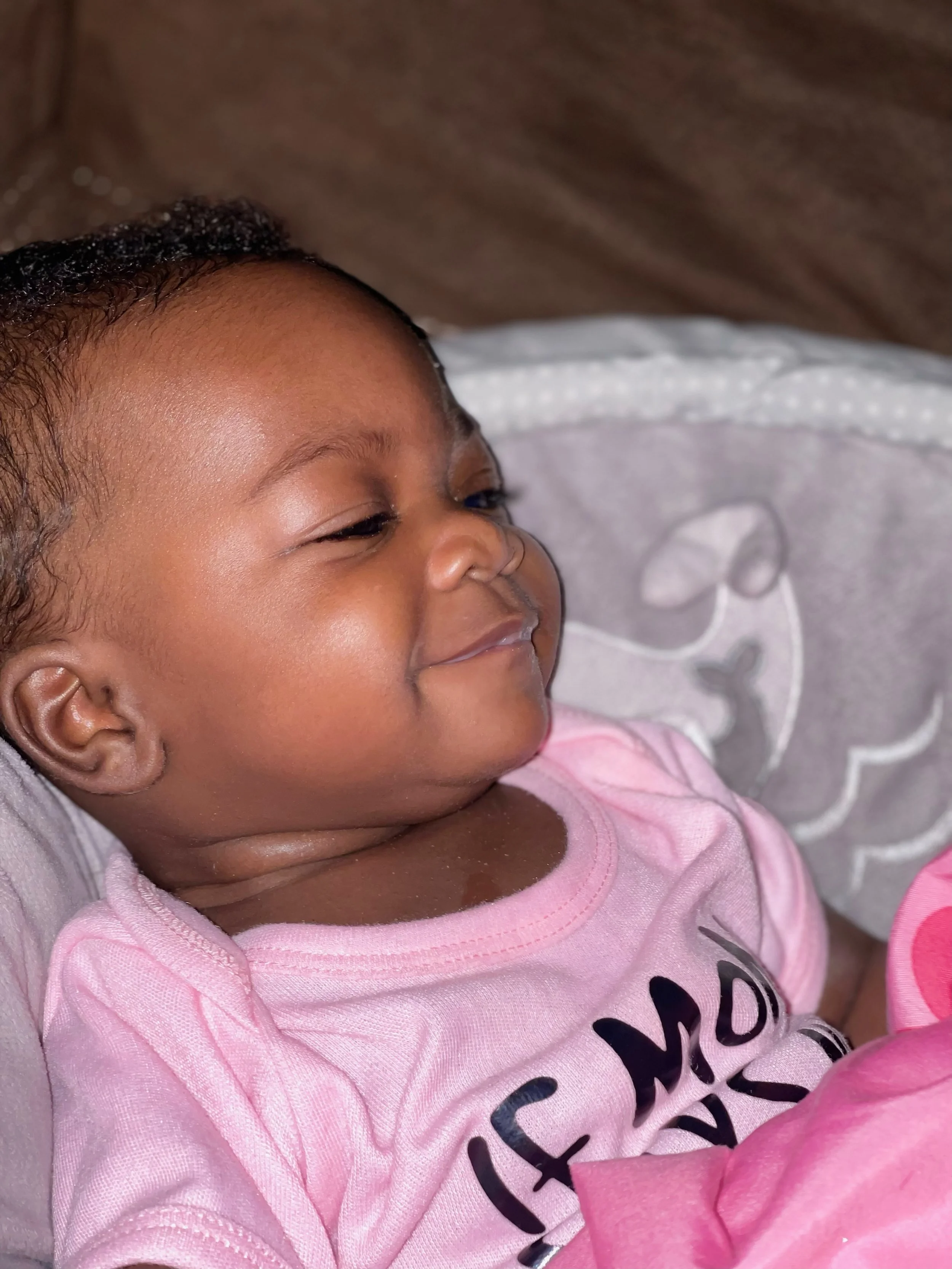Close-up of a smiling infant lying on a gray blanket with a white pattern, wearing a pink shirt.