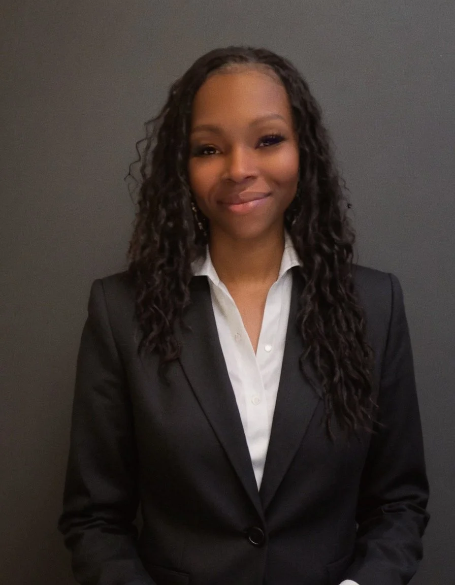 A young African American woman with curly hair wearing a black blazer and white shirt, smiling against a dark background.