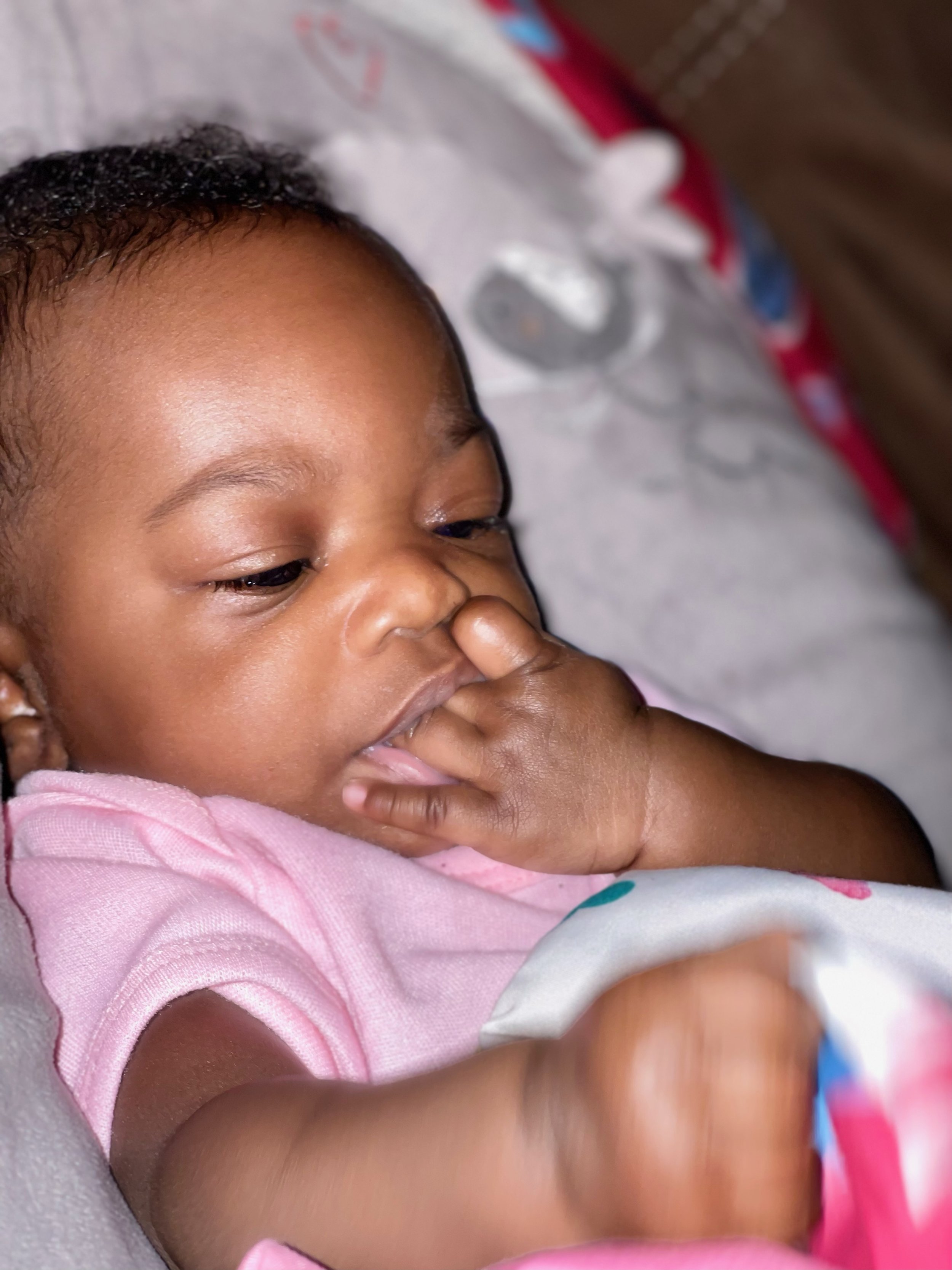 Close-up of a young child with short dark hair and brown skin, lying down and wearing a pink shirt, with a hand near their nose and mouth.