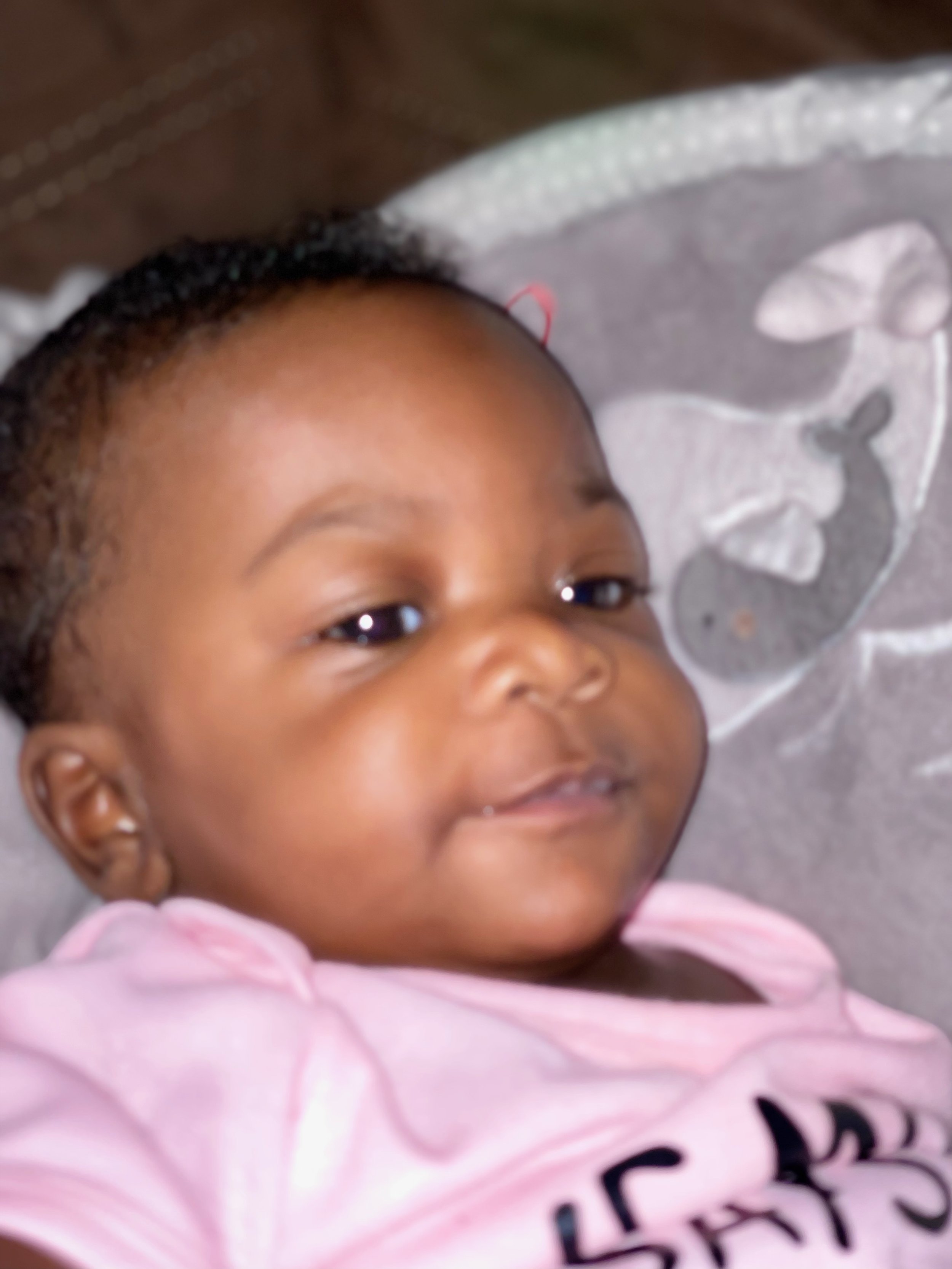 Close-up of a baby lying down, wearing a pink shirt, with a cheerful expression.
