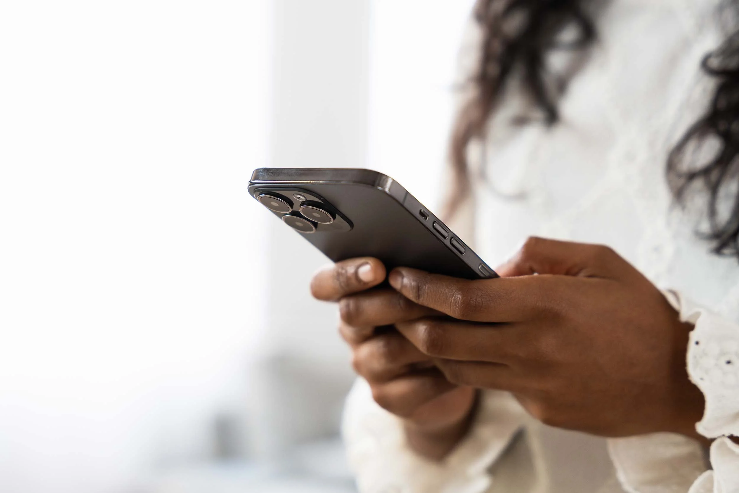 Close-up of a person with dark skin using a smartphone with three camera lenses on the back, handheld in front of a white background.
