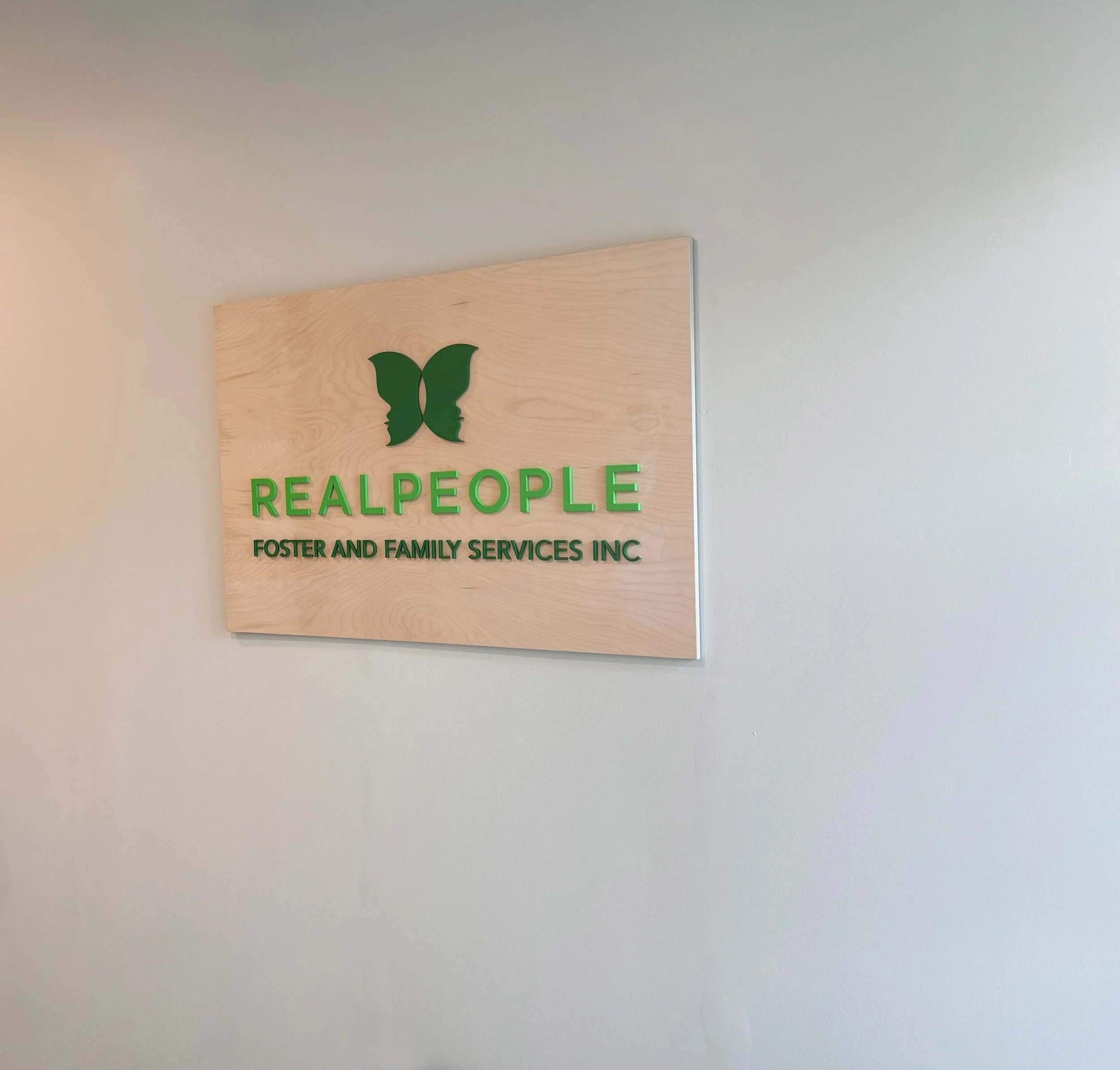 Wooden sign on a white wall displays the logo and name of RealPeople Foster and Family Services Inc, with a green butterfly logo and green text.
