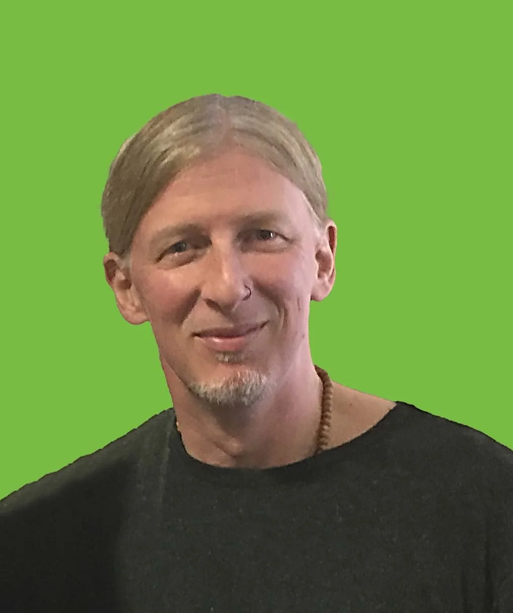 Portrait of a smiling man with light brown hair, a goatee, wearing a black shirt, and a beaded necklace, against a green background.