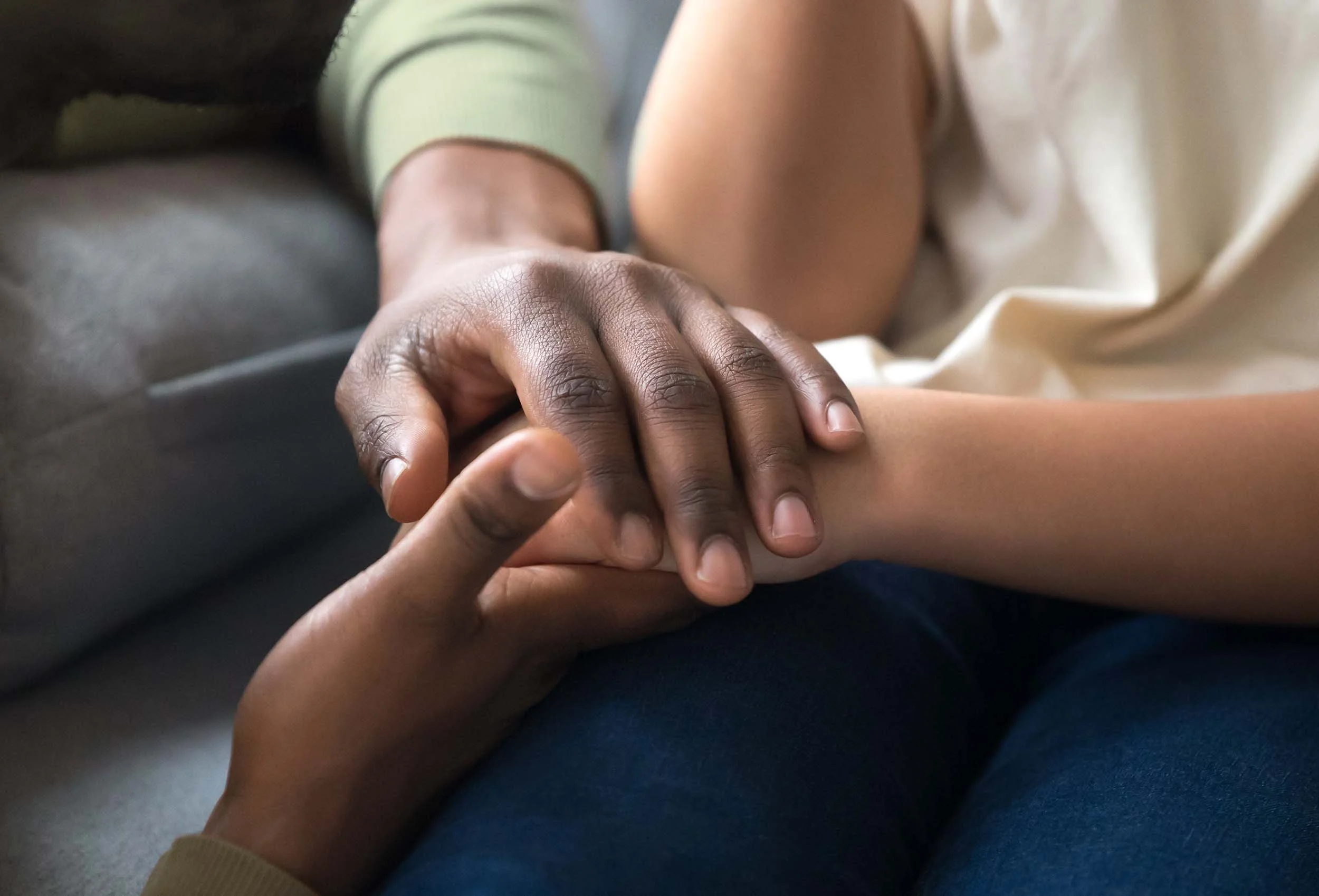 Close-up of two people holding hands, one with darker skin and the other with lighter skin, sitting close together on a couch.