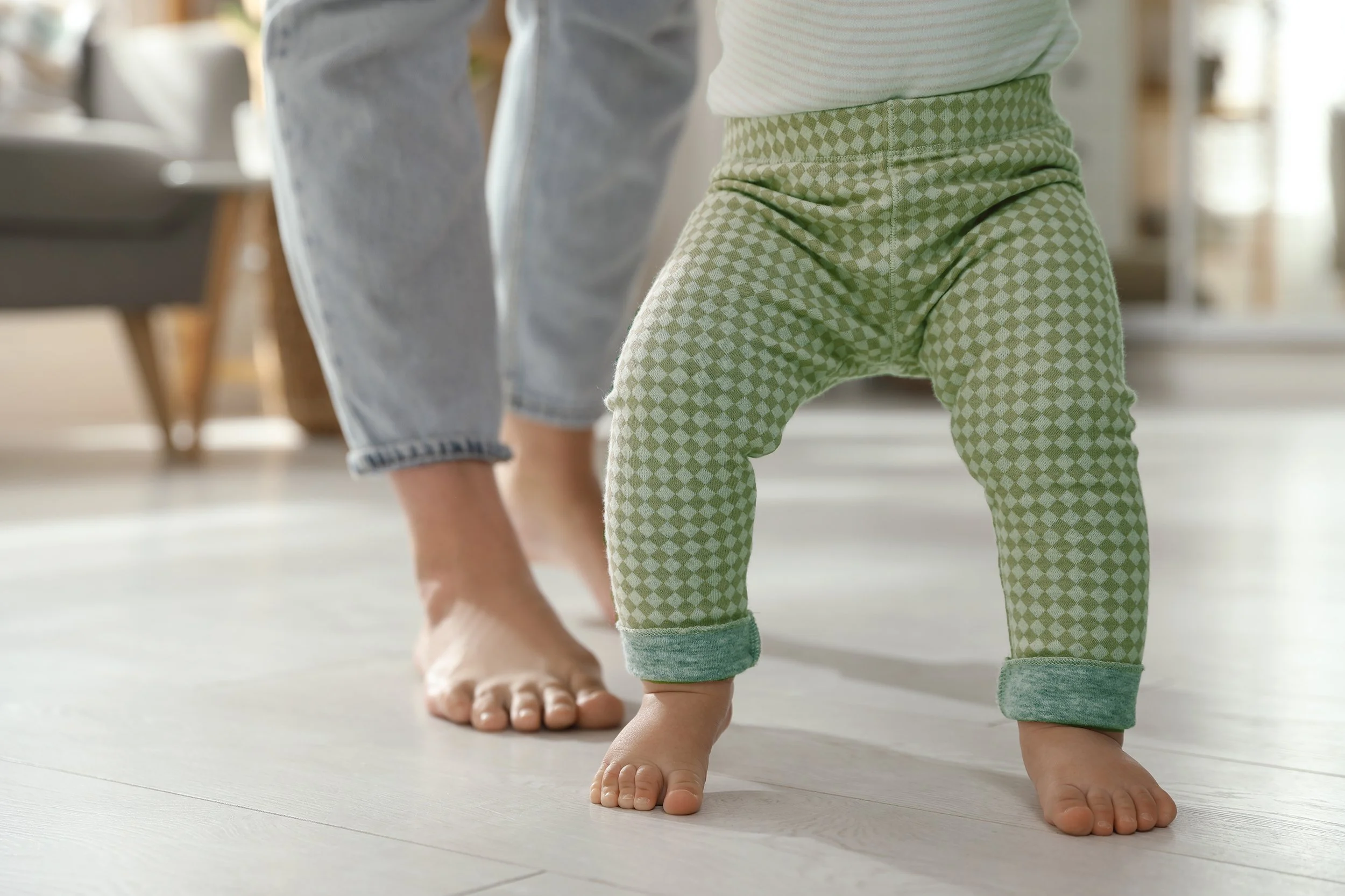 Close-up of a toddler's legs and feet as they crawl on a light-colored wooden floor, wearing green checkered pants and gray socks, with an adult's feet and legs visible in the background.