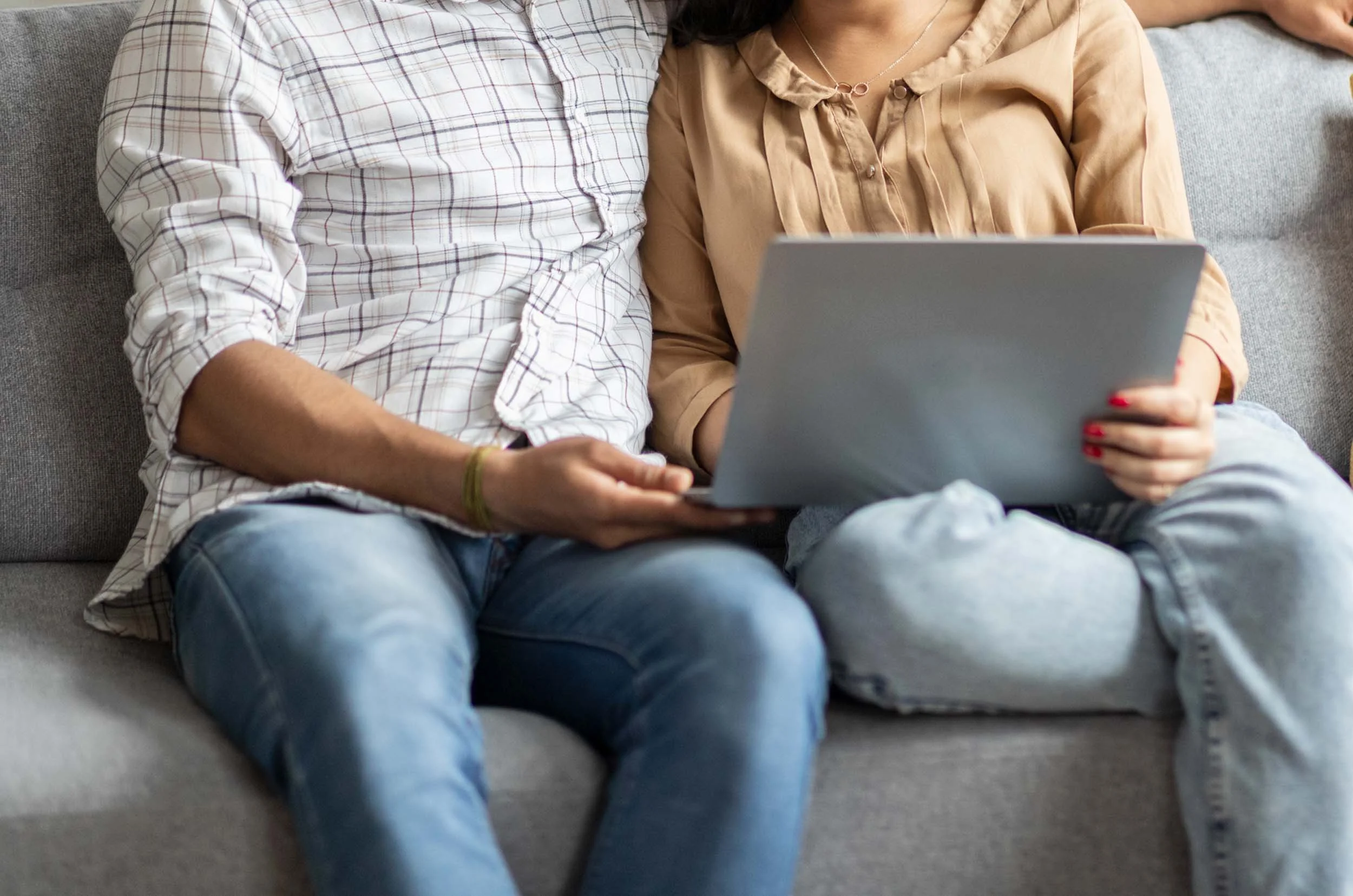 Two people sitting on a gray sofa, looking at a gray laptop together, with one person's arm around the other.