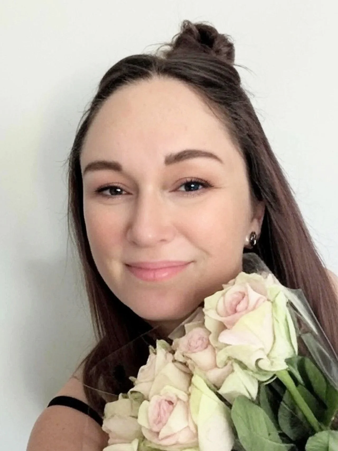 A young woman with long brown hair styled with a small bun, holding a bouquet of pale pink and white roses, and smiling softly at the camera against a plain white background.