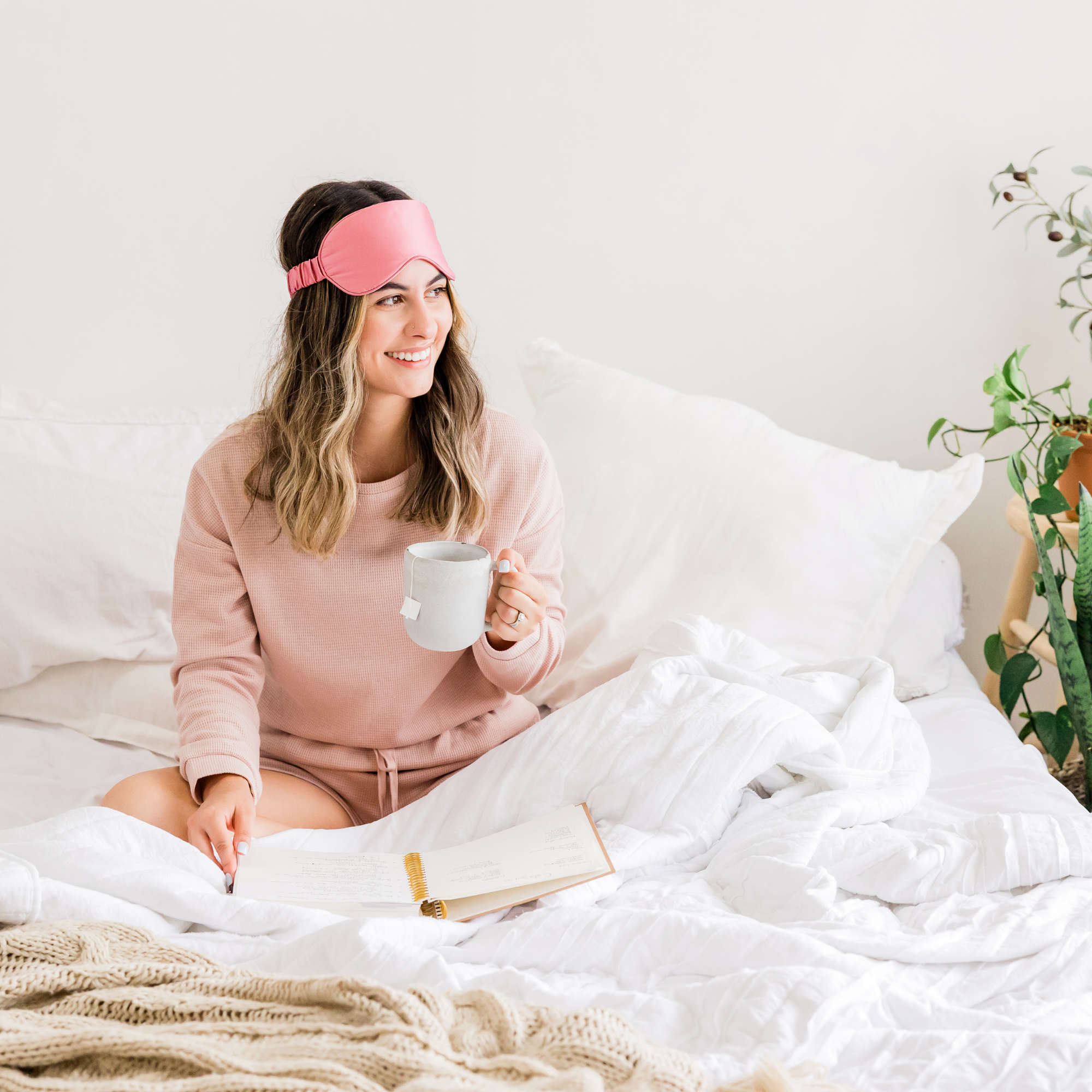 Woman sitting in bed with pink sleep mask on her forehead, holding a gray mug, reading a book, surrounded by white bedding and pillows, with a green plant on the side.