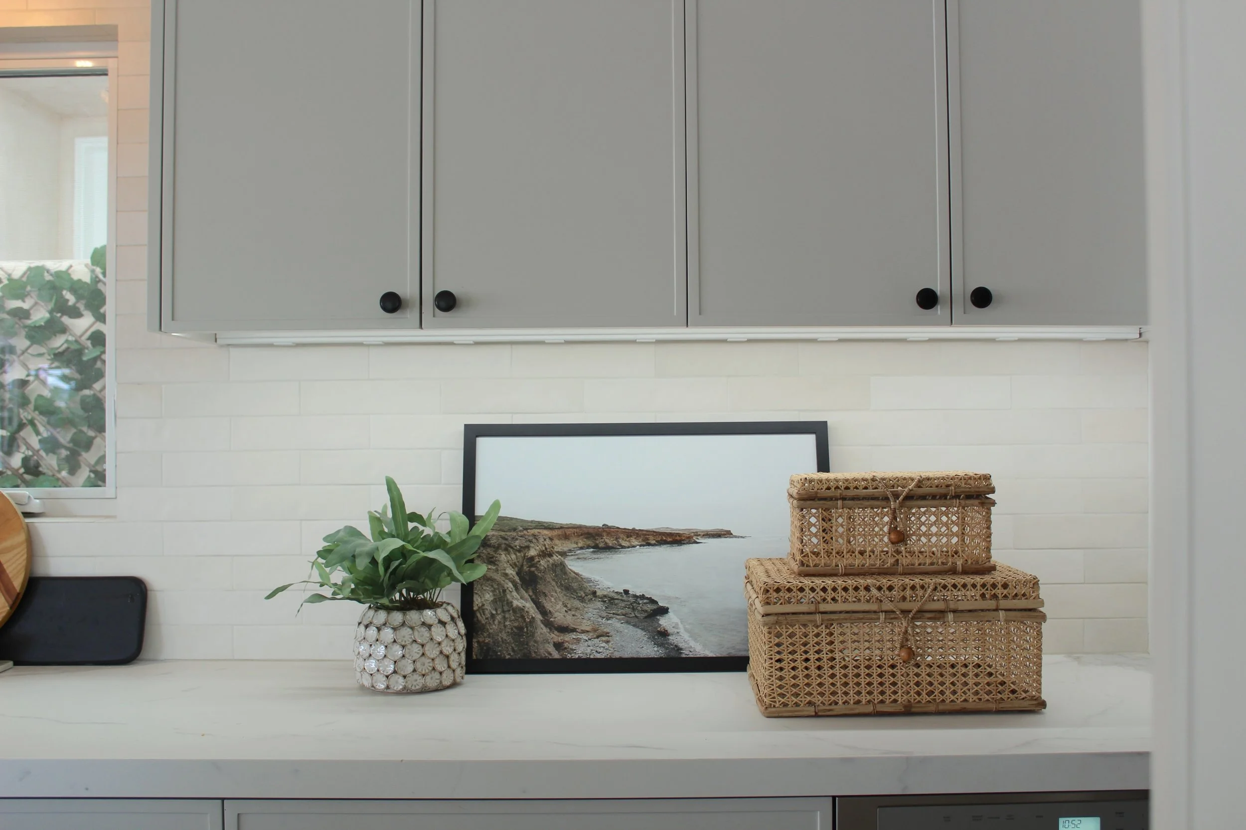 Home decor with a potted plant, framed picture of a coast, and two stacked wicker baskets on a white kitchen counter, with gray cabinets above.
