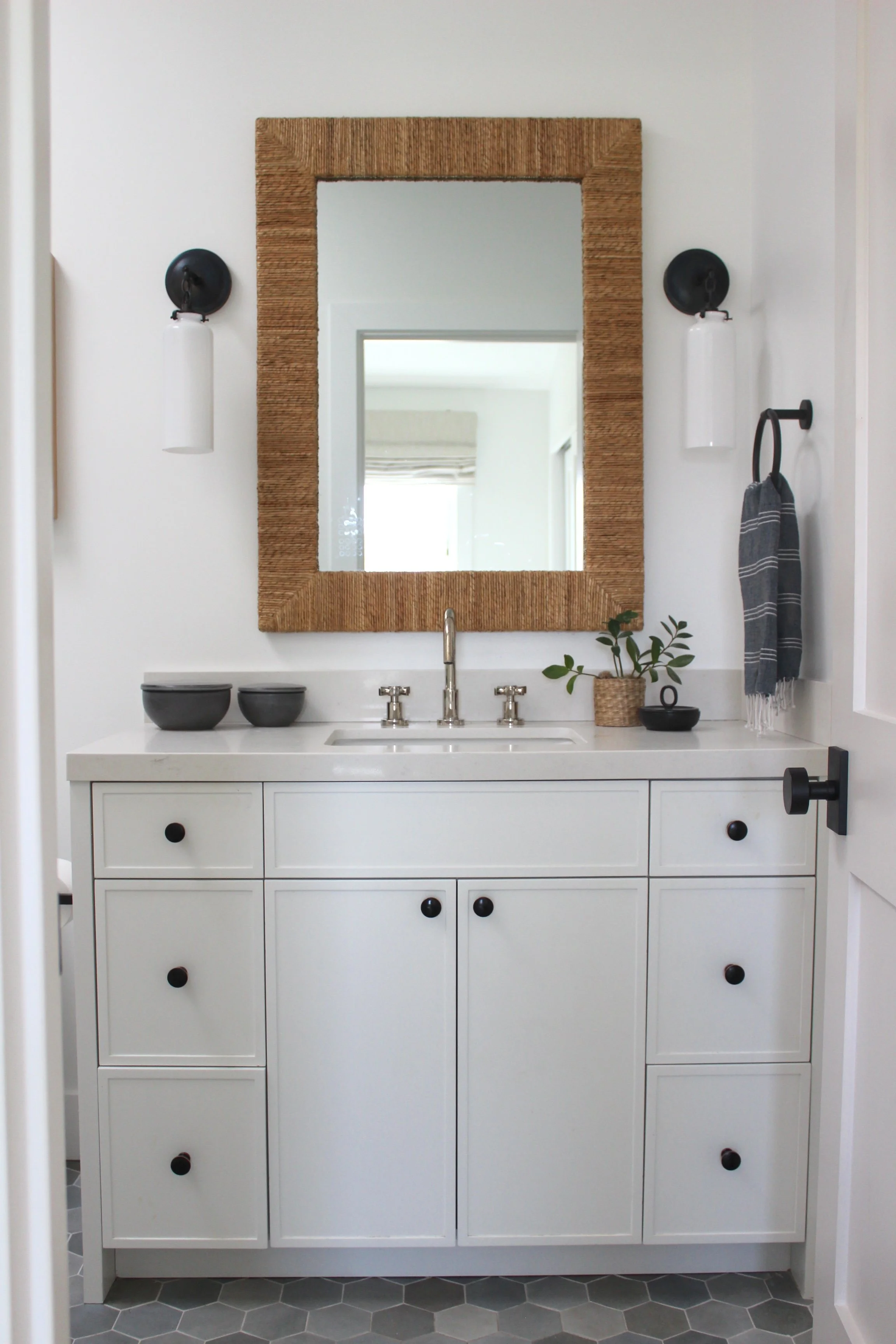Modern bathroom vanity with white cabinets, a mirror with a woven wood frame, black knobs, black and white accessories, and a small plant on the countertop.