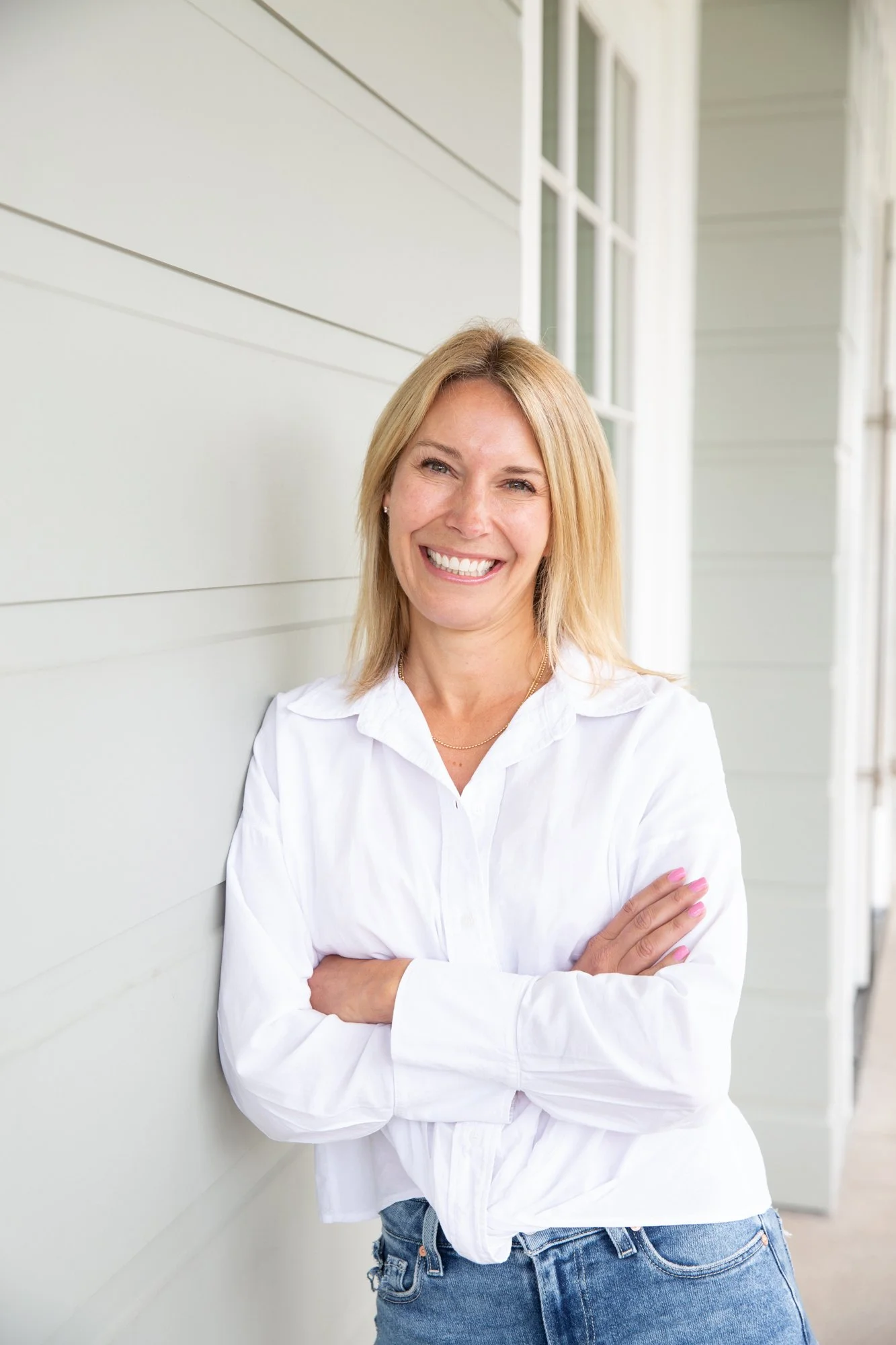 Portrait of a smiling blonde woman with arms crossed, standing outdoors against a light-colored wall with windows.