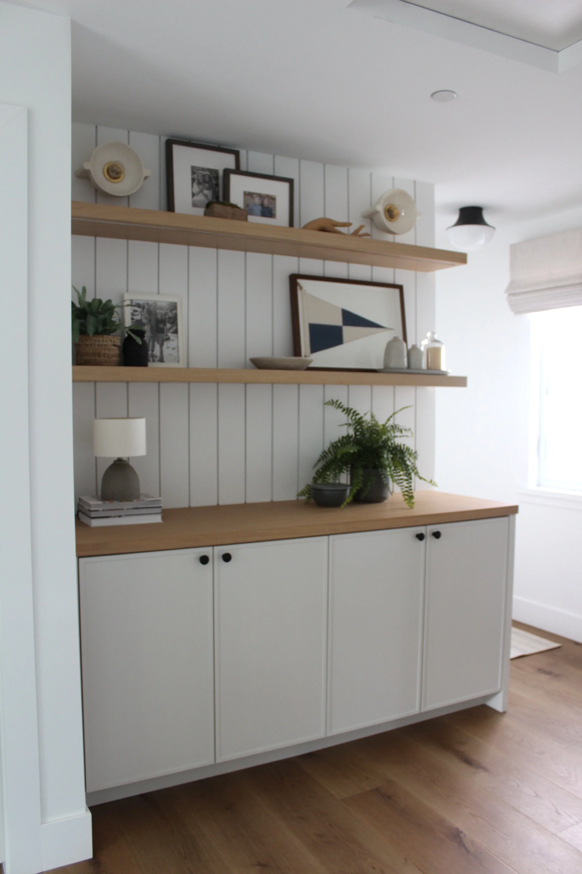 Interior of a living room corner with two wooden floating shelves holding framed photos, decor, and a small sculpture, above a white cabinet with black knobs, a potted plant, and decorative items on the wooden surface.