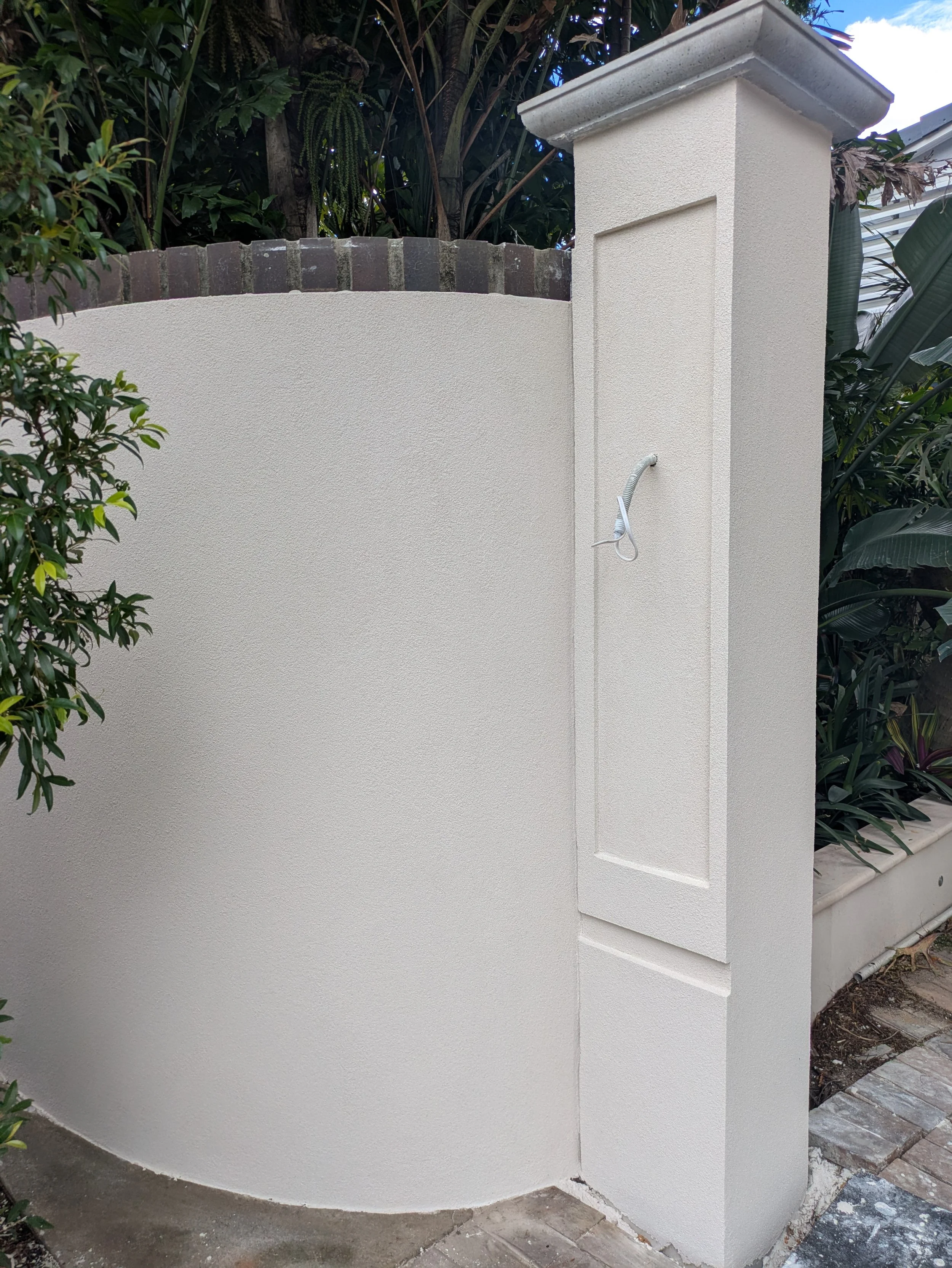 Exterior corner of a white stucco wall with a decorative post and greenery in the background.