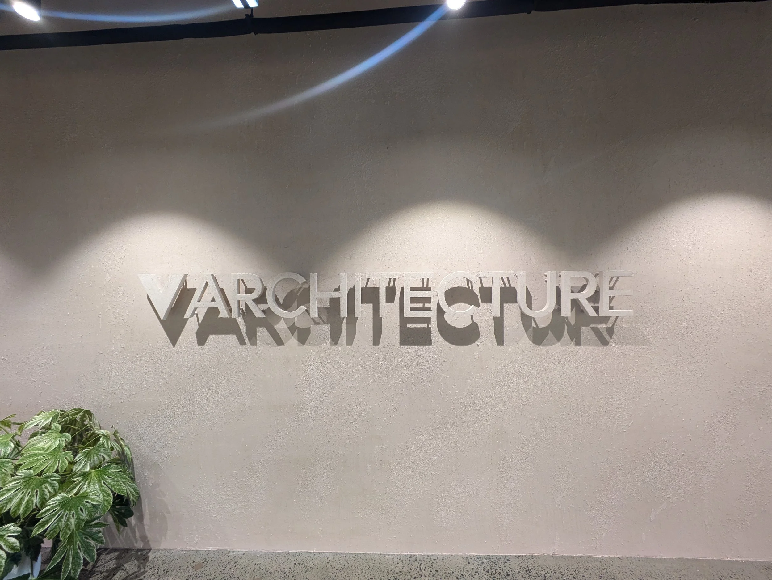 The image shows a wall with the text 'VARCHITECTURE' in metallic letters, illuminated by three ceiling spotlights, with a plant at the bottom left.