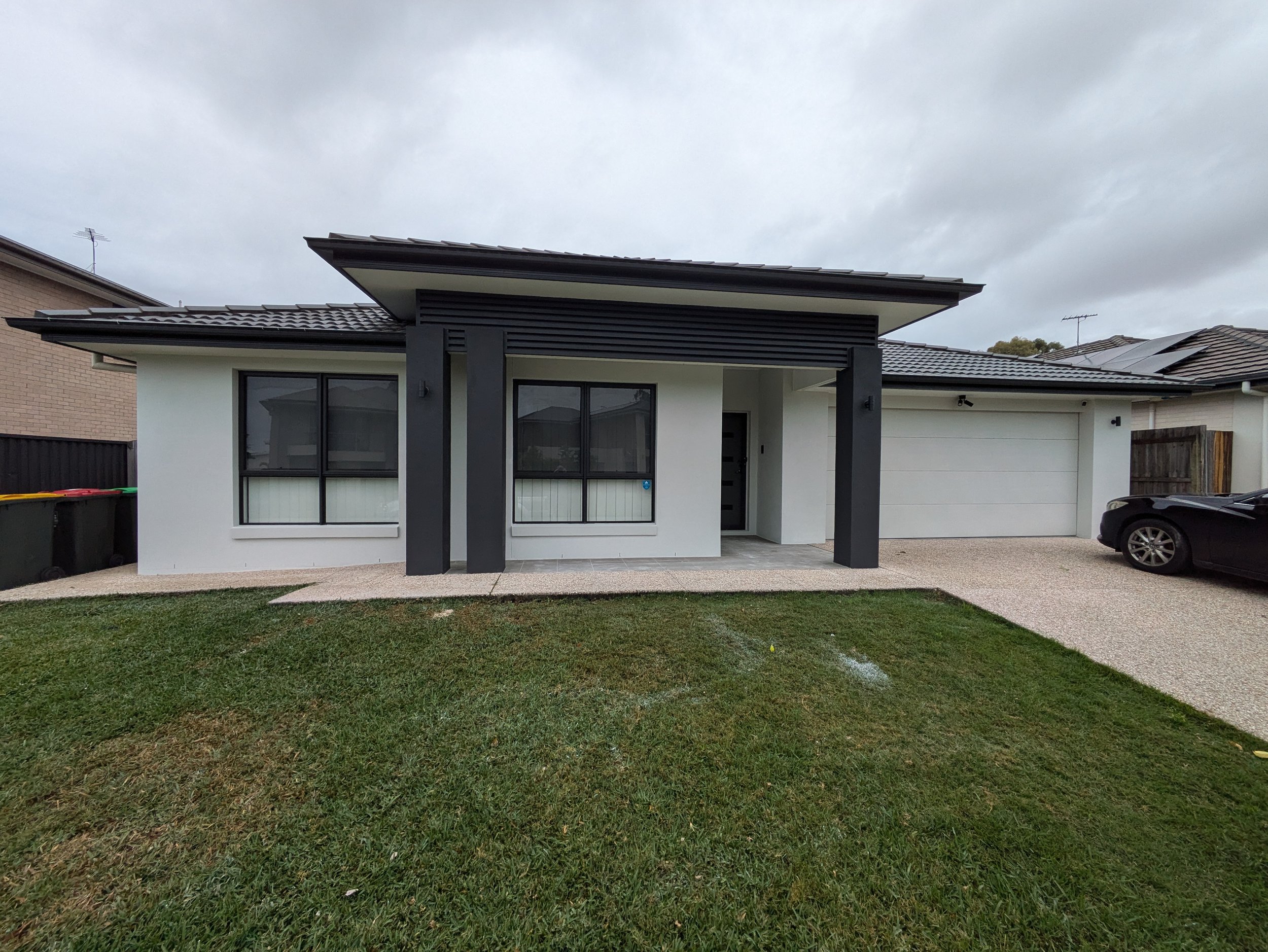 Modern house with white walls, black window frames, and a black garage door, with a car parked in the driveway.