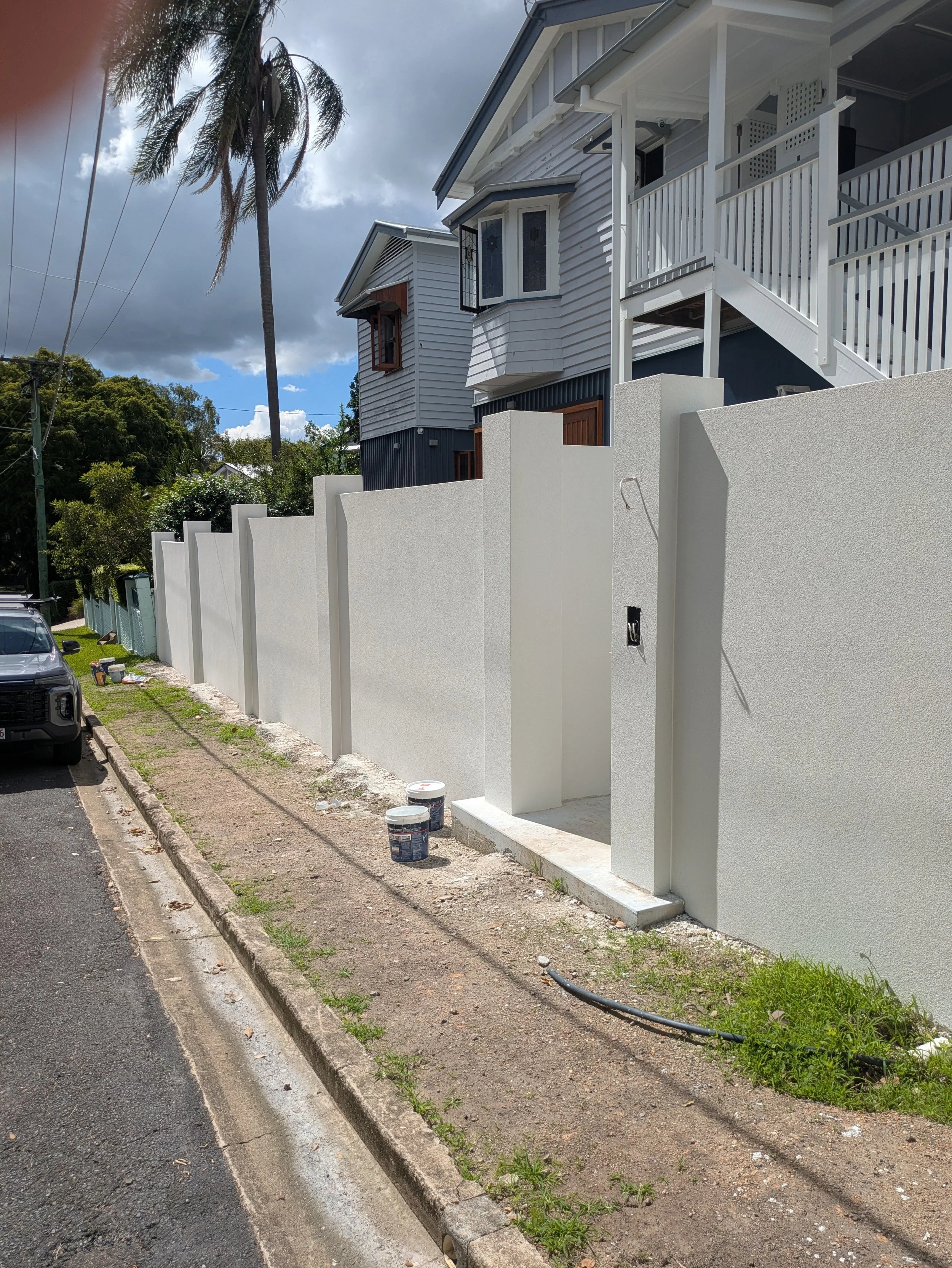 A white concrete wall with a gate under construction along a sidewalk, with buckets of paint or plaster and a pipe lying on the ground, and residential houses in the background under a partly cloudy sky.