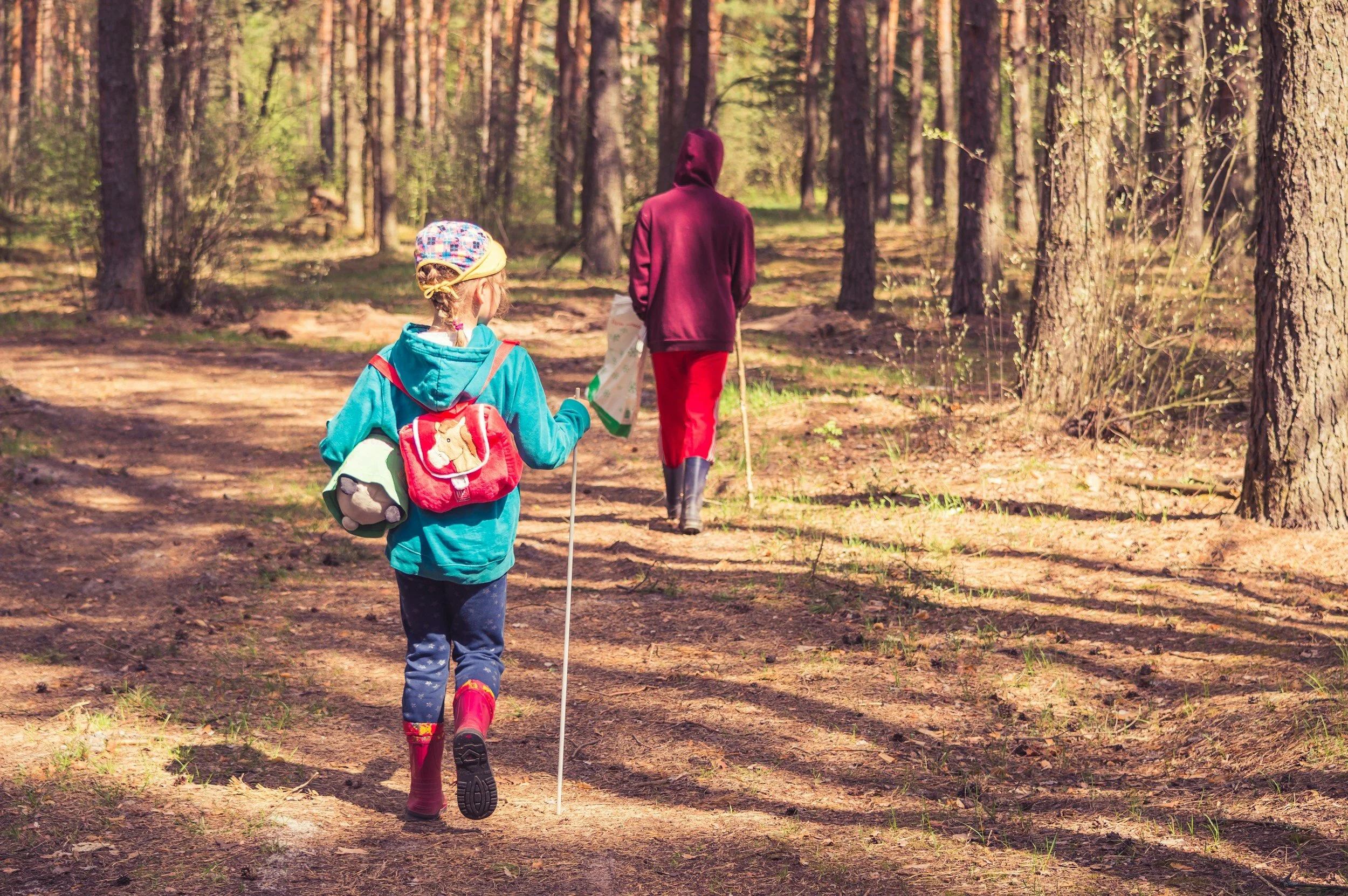A young girl and an adult walking on a forest trail. The girl is wearing a colorful hat, blue jacket, and rain boots, while the adult is dressed in a maroon hoodie, red pants, and rain boots, carrying a walking stick.
