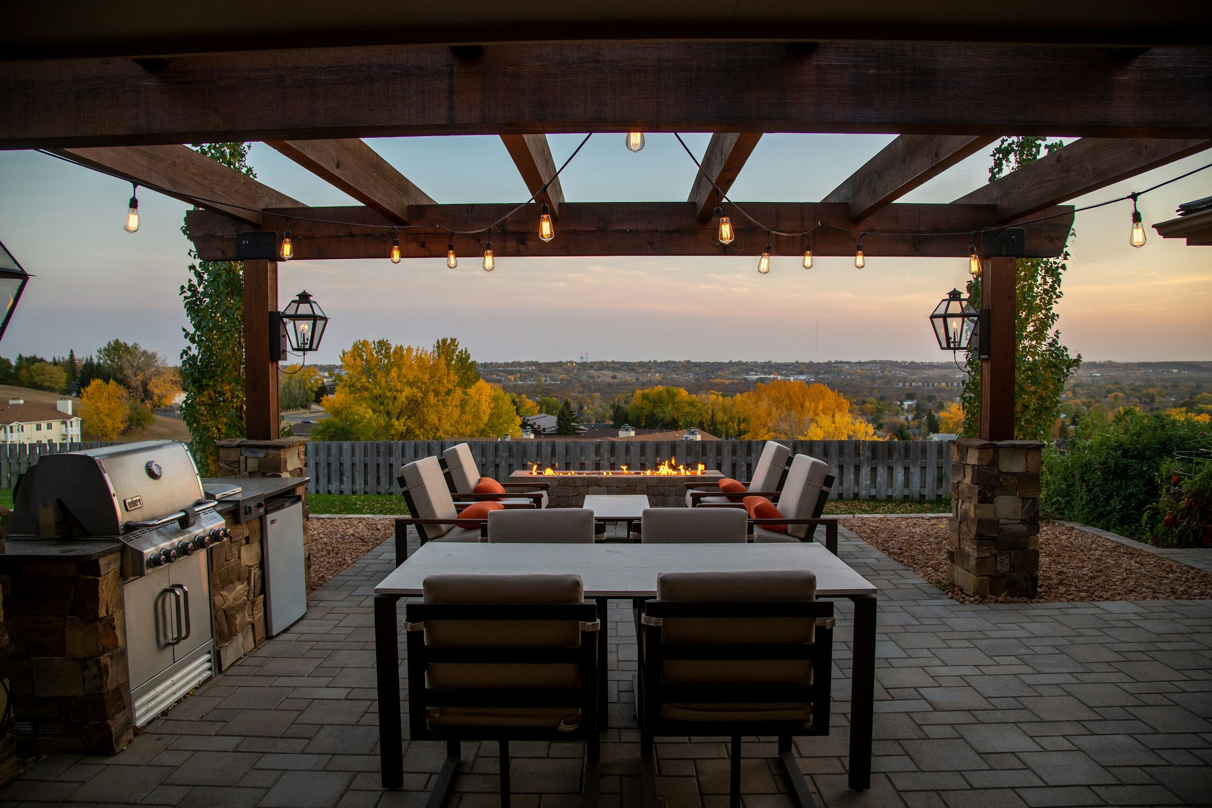A backyard patio with seating, a fire pit, a grill, string lights overhead, and a view of autumn trees and a distant landscape at sunset.