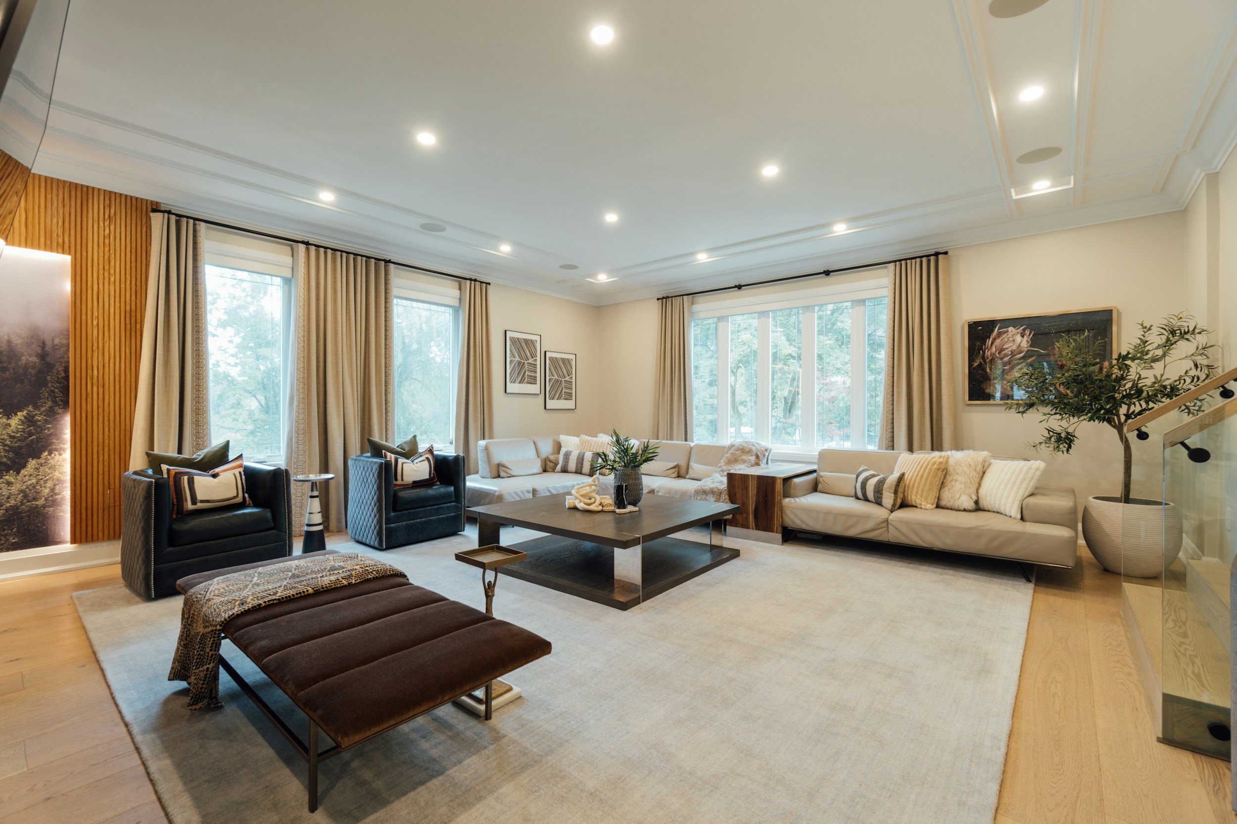 A spacious living room with large windows, beige curtains, a white sectional sofa, black and gray armchairs, a wooden coffee table, a potted plant, wall art, and a light-colored rug.