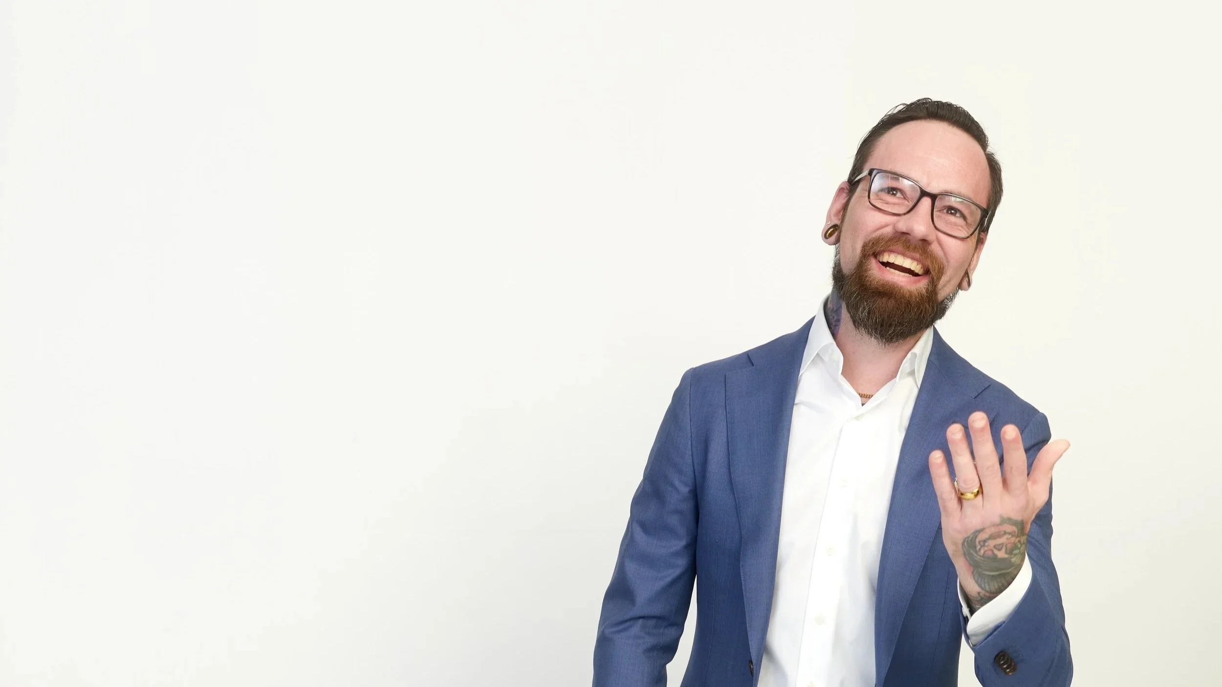 A man with glasses, a beard, and tattoos is smiling and pointing towards the camera. He is wearing a blue suit and a white shirt against a plain white background.
