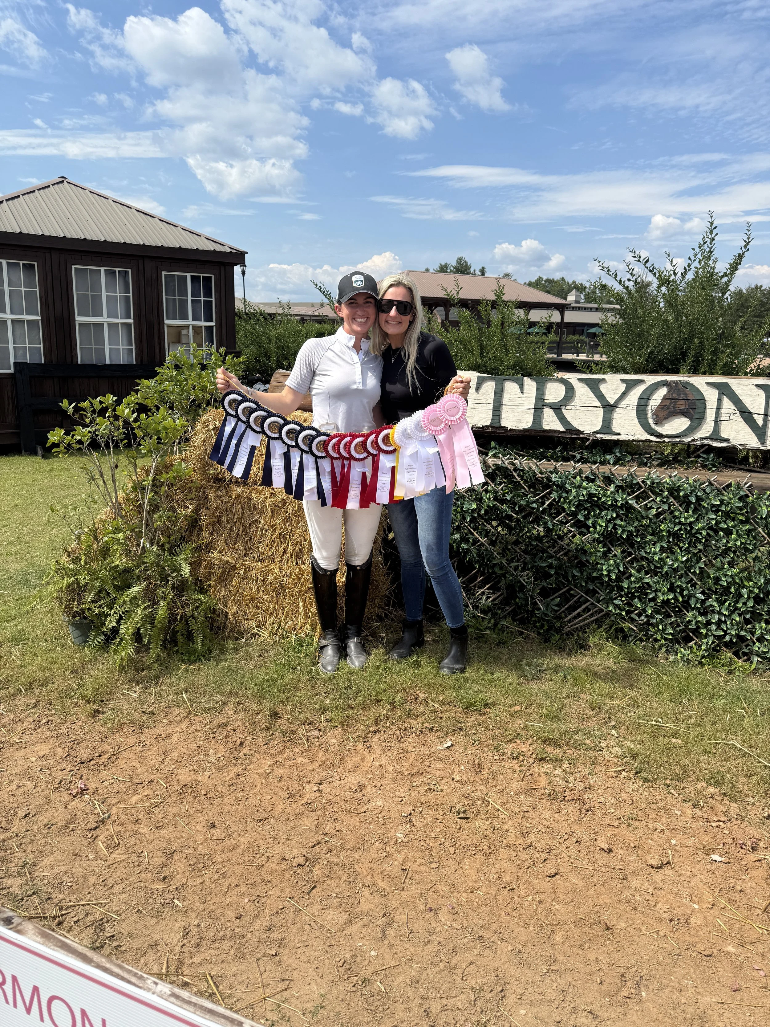 Two women standing outdoors, smiling, holding a sash of award ribbons. One woman is wearing a white riding outfit with a black helmet, and the other is dressed in a black top, jeans, and sunglasses. They are next to a hay bale with greenery and a woo