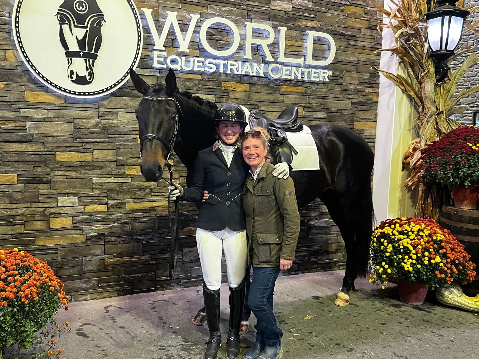 Two women standing in front of a black horse with a saddle at the World Equestrian Center, smiling and embracing each other. One woman is dressed in equestrian riding attire, including a helmet, riding jacket, and white pants, while the other is dres