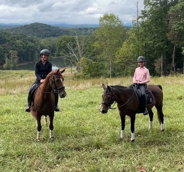 Two people riding horses in a grassy field with a scenic background of trees, hills, and a cloudy sky.