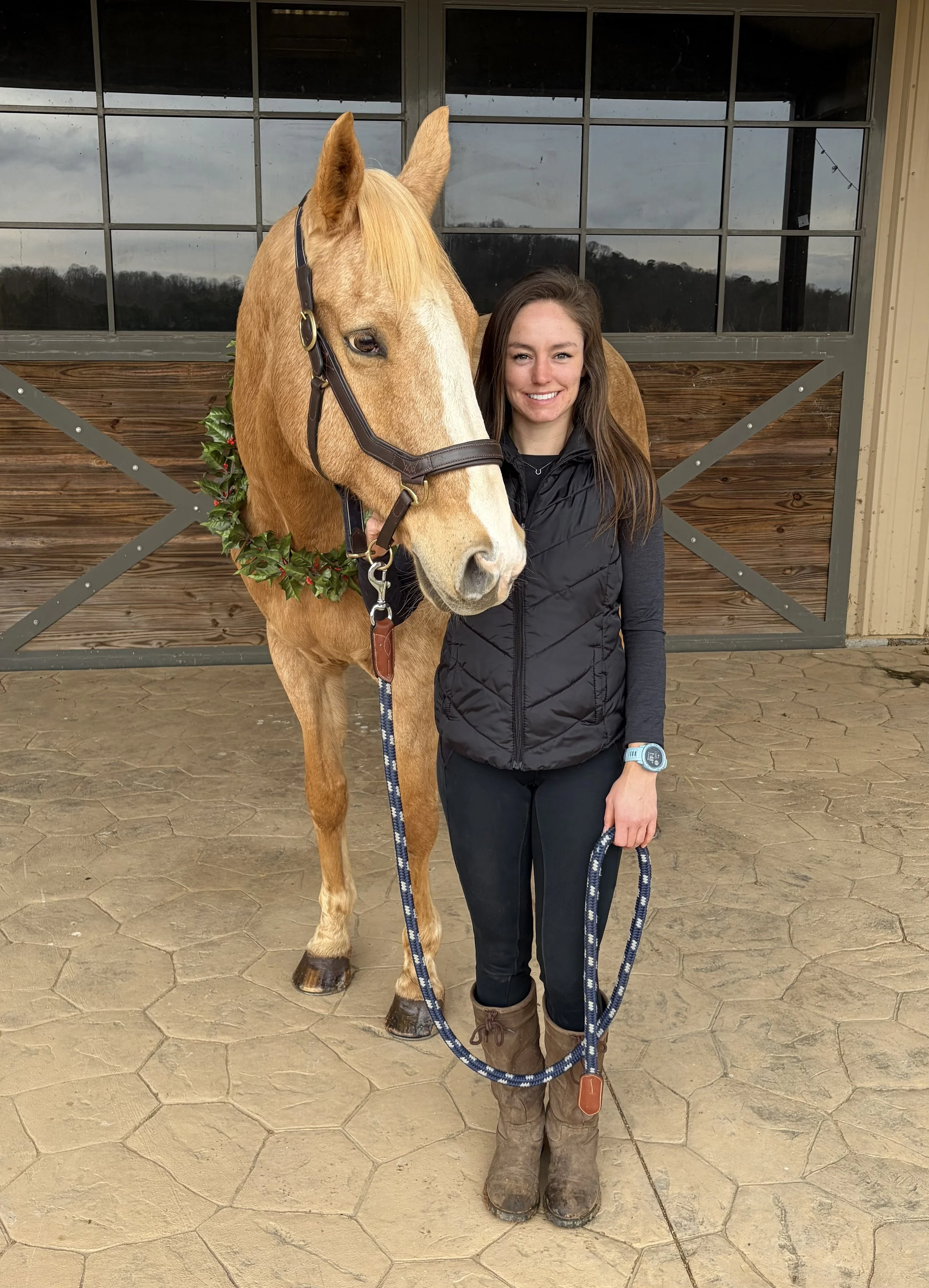 A young woman standing next to a light brown horse with a white blaze on its face, in front of a stable door. The horse is wearing a dark halter and a decorative wreath around its neck, and the woman is smiling, holding the horse's lead rope.