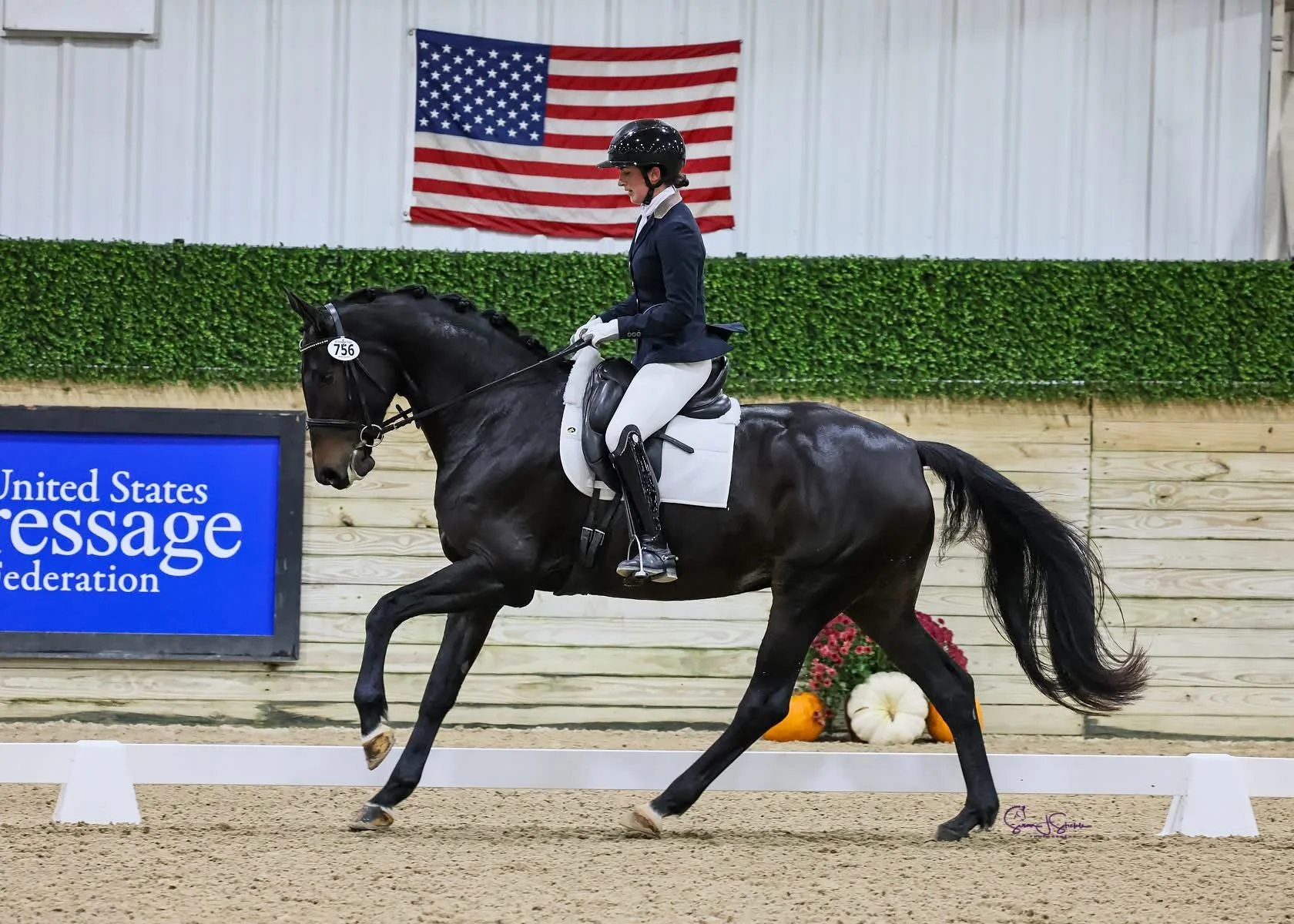 A female equestrian dressed in formal riding attire, including a black helmet and white gloves, riding a dark brown horse with a braided mane in an indoor riding arena. The arena features a wooden fence with pumpkins and flowers, an American flag han