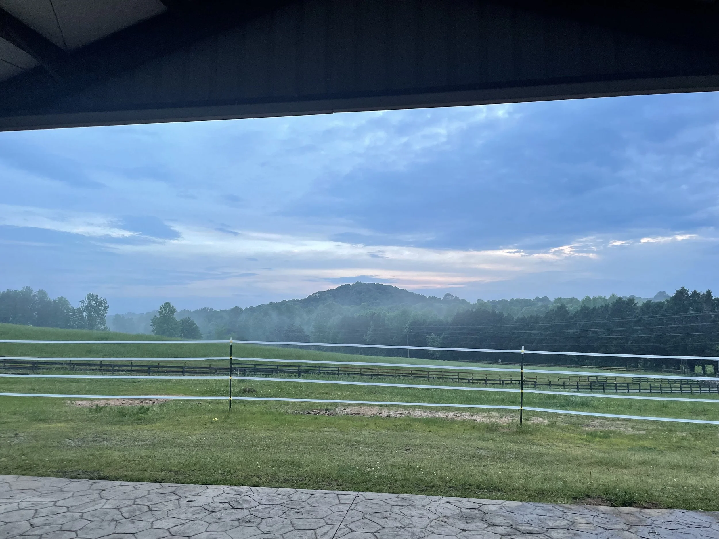 View of a rural landscape with rolling green hills, trees, and a distant mountain, seen through a window with a dark overhead beam. The sky is partly cloudy at dusk.