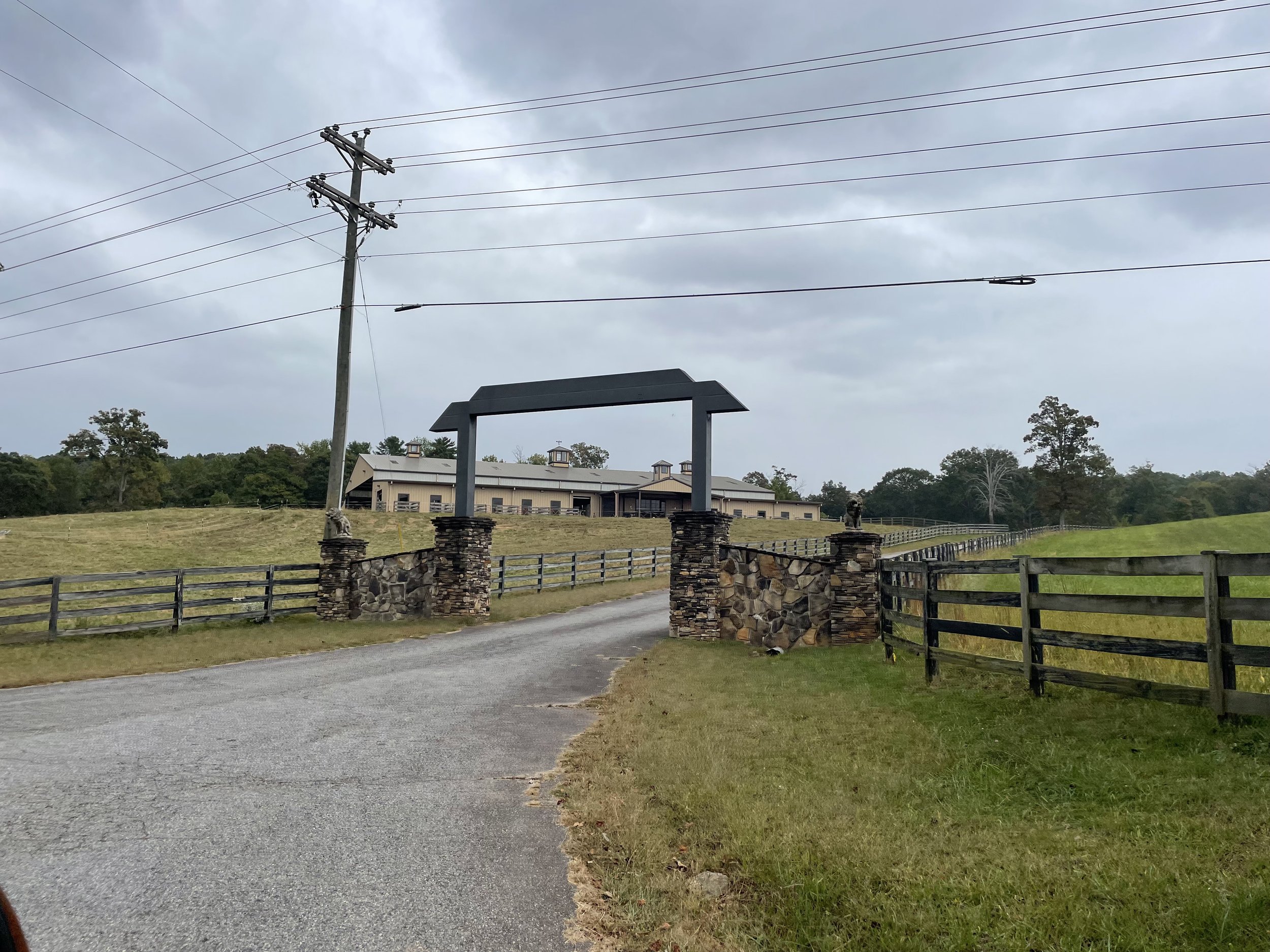 Entrance gate to a property with a stone and wood archway, a gravel driveway, fenced green fields, and a large beige building in the background under a cloudy sky.