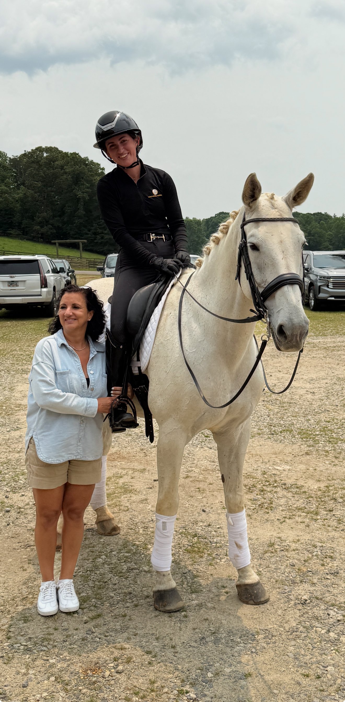 A woman dressed in black riding a white horse with a woman in a light blue shirt and beige shorts standing beside them.