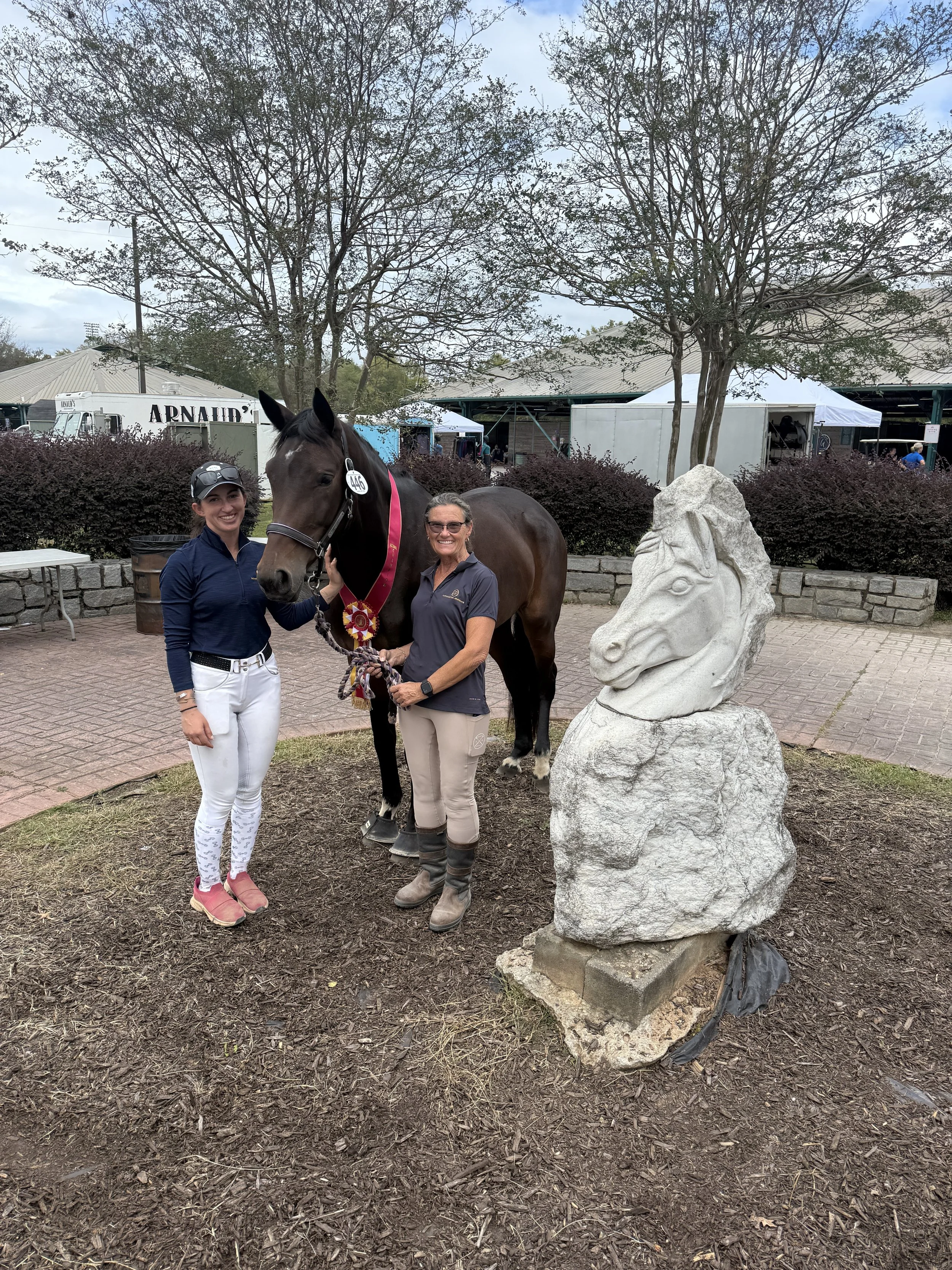 Two women standing beside a dark brown horse with a pink ribbon and rosette, at a horse show. One woman is holding the horse's bridle and the other is smiling. A stone horse head sculpture is nearby, and trees and event booths are in the background.