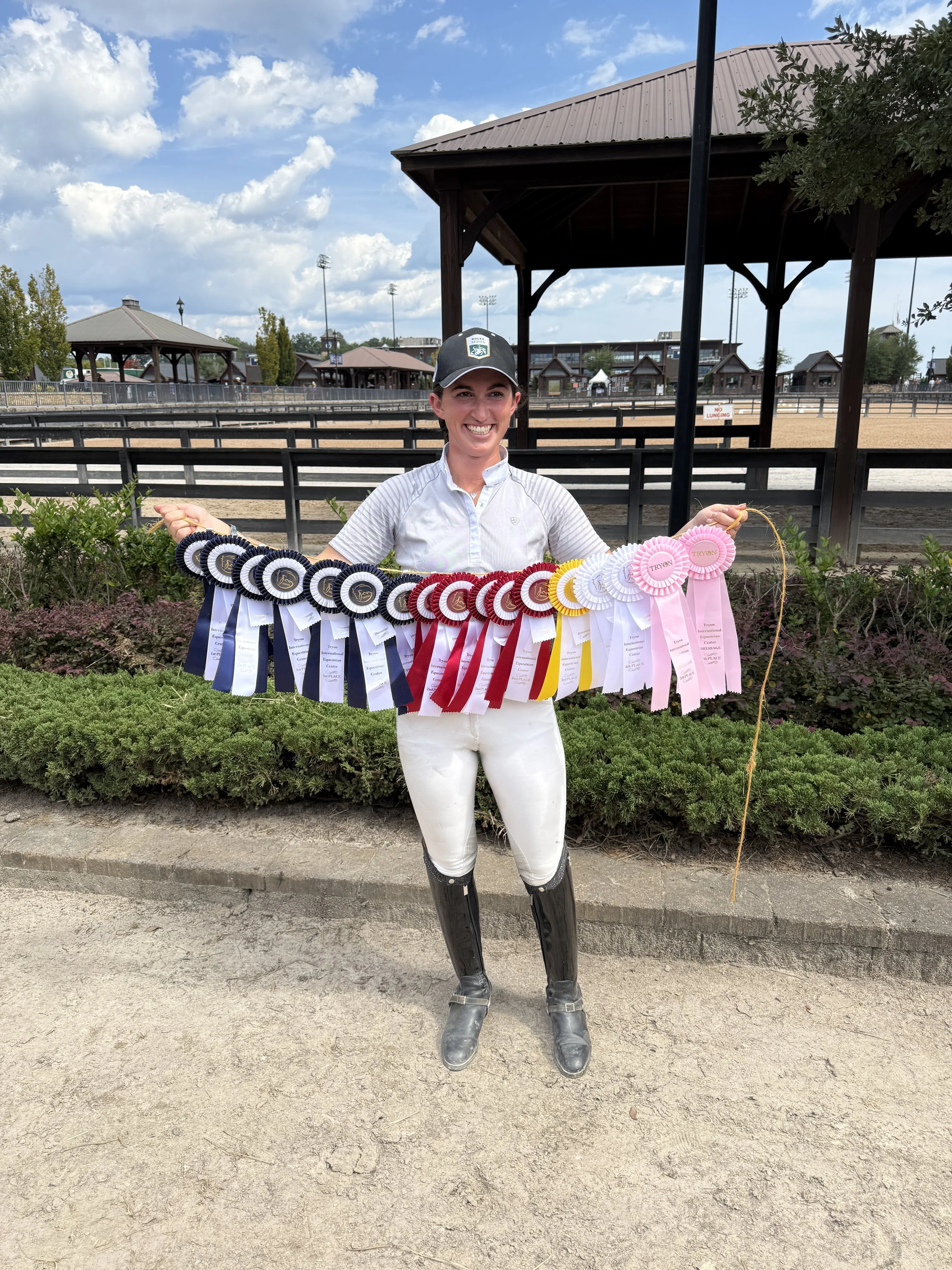 A smiling woman in equestrian riding gear standing in front of a horse arena, holding a colorful display of riding competition ribbons.