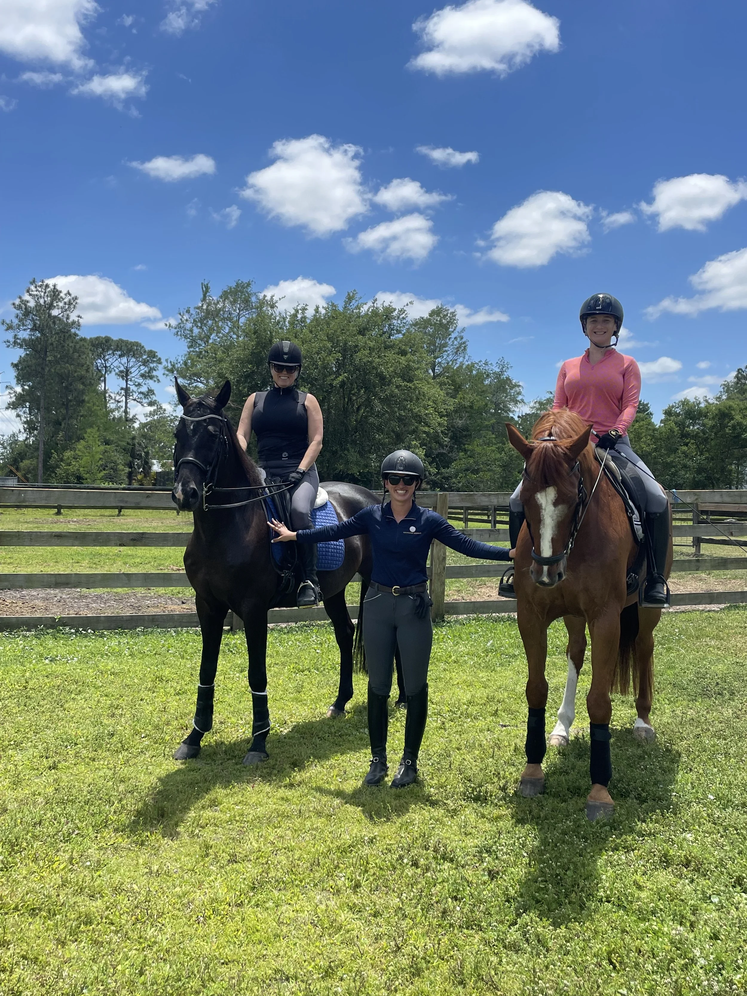 Two women riding horses and a woman standing on the ground between them, all wearing riding helmets, in an outdoor grassy riding area with a wooden fence and green trees under a partly cloudy blue sky.