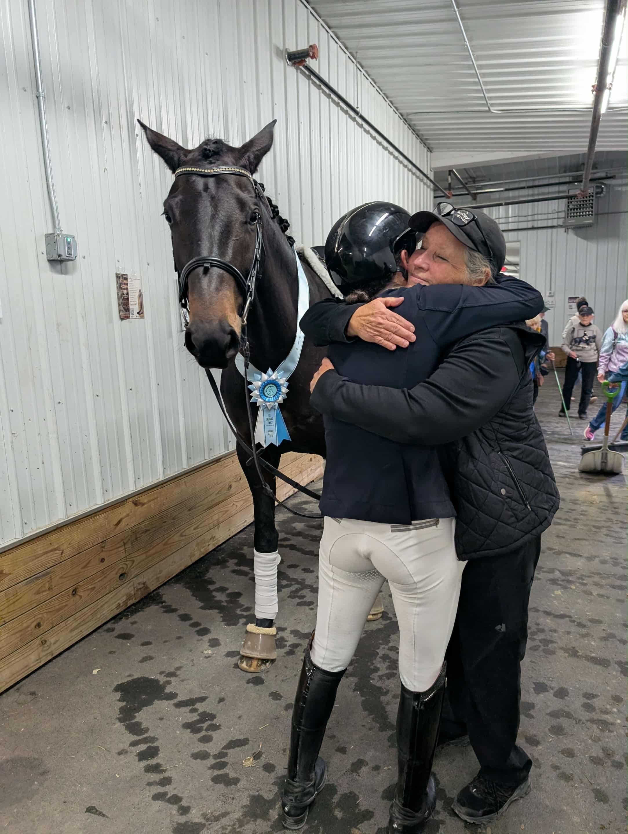 A woman and a man hug in a barn, with a black horse wearing a blue ribbon and a white sash standing next to them. The woman is wearing riding gear, including tall black boots, beige pants, and a black jacket. There are other people and barn equipment