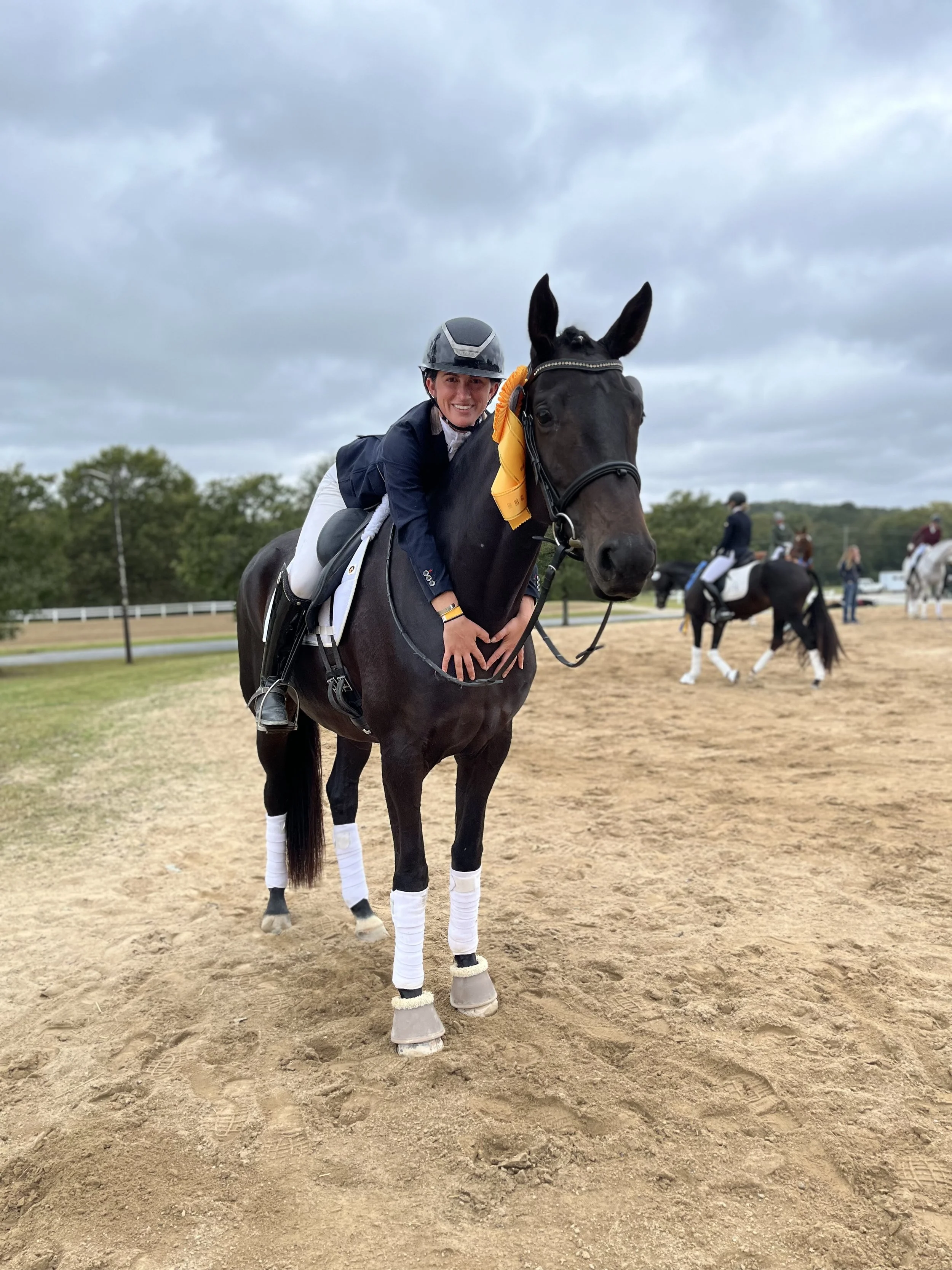 A smiling woman in equestrian gear riding a black horse with a yellow ribbon on its bridle, standing on a sandy riding arena with other riders and horses in the background under cloudy skies.