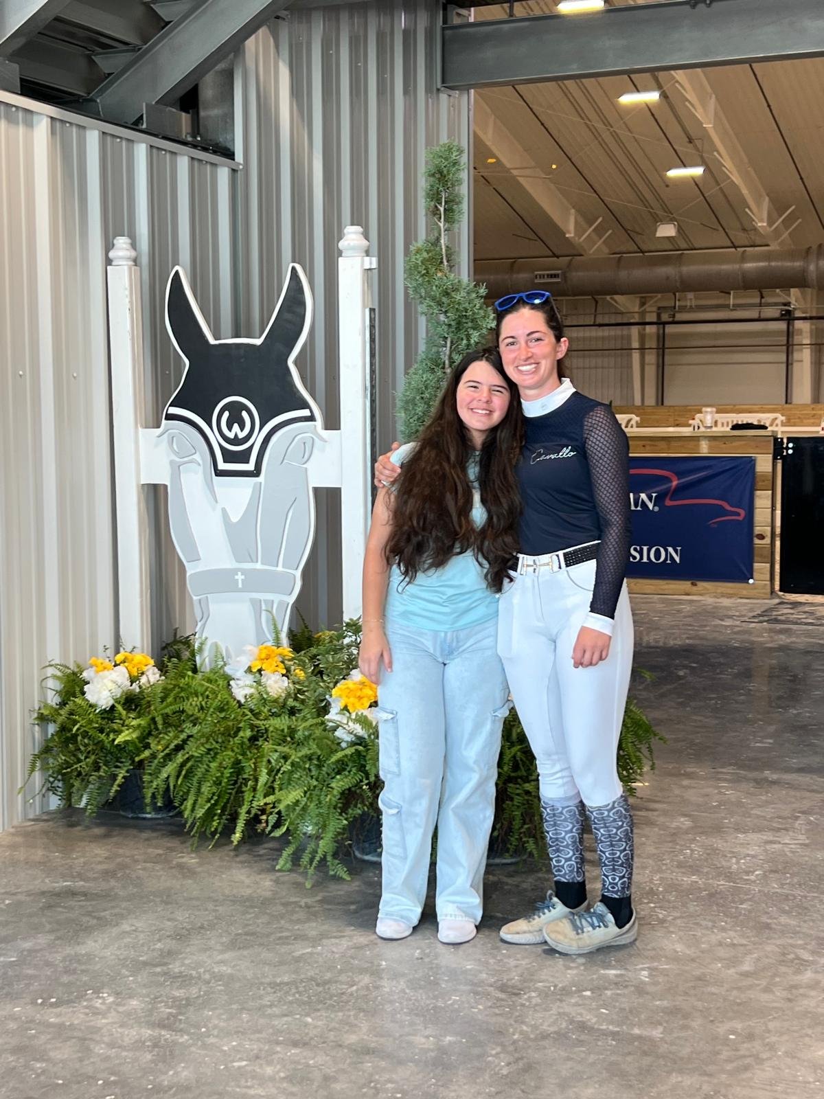 Two women standing close together, smiling, in front of a donkey-themed decoration with flowers at their feet, inside a large industrial-style building.