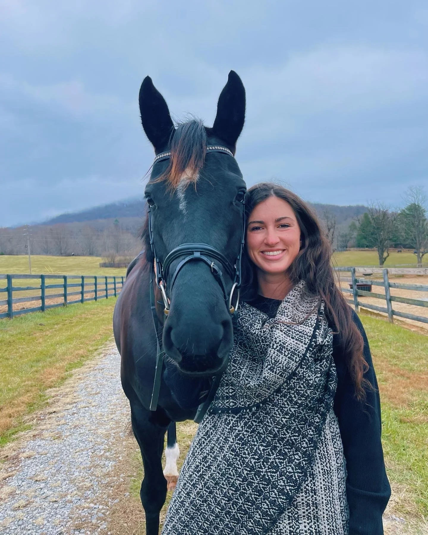 A young woman standing next to a black horse on a farm, smiling at the camera, with fences and trees in the background on a cloudy day.