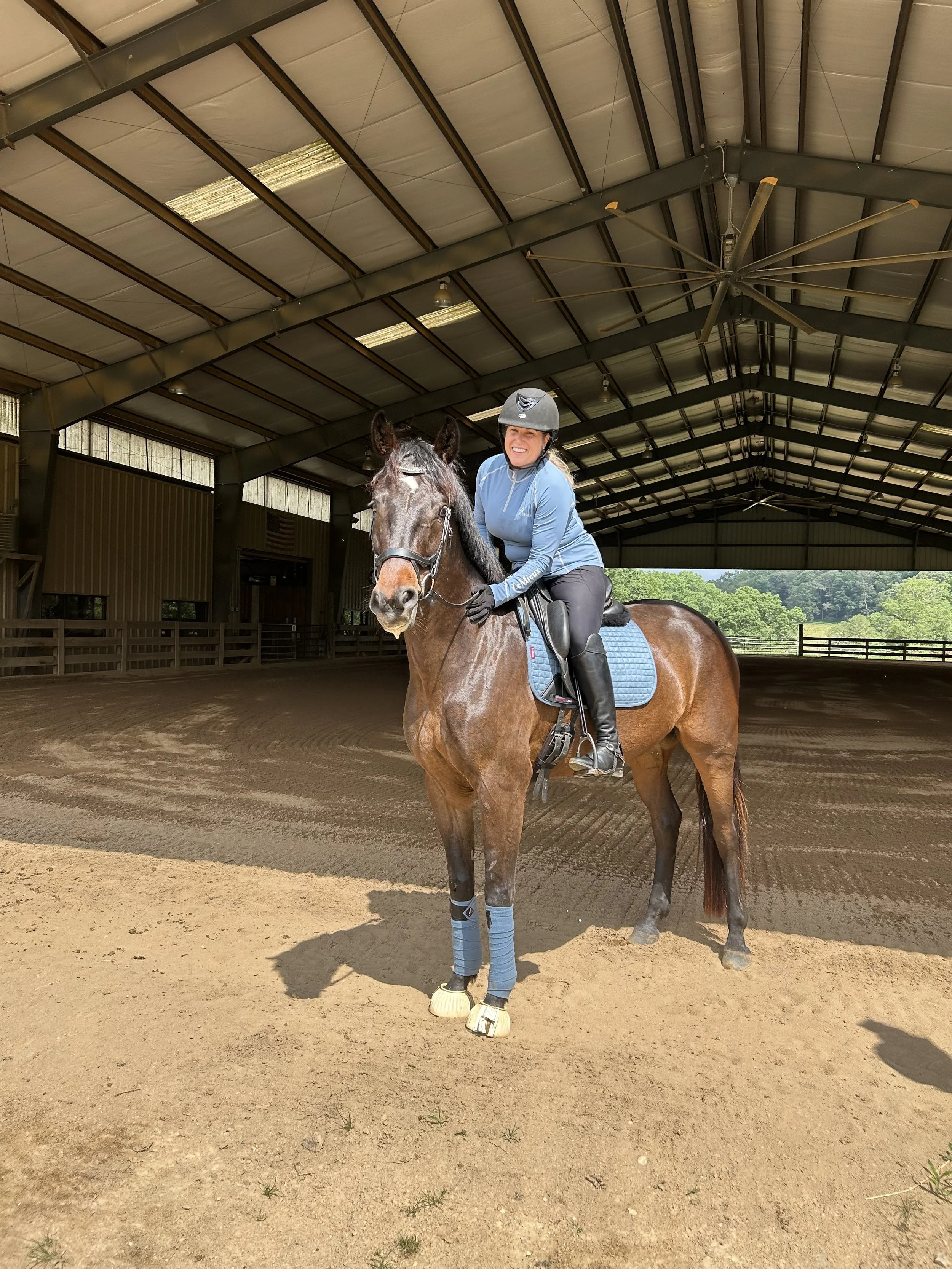 A woman in equestrian gear riding a brown horse inside an indoor riding arena with a high ceiling and large ceiling fan.
