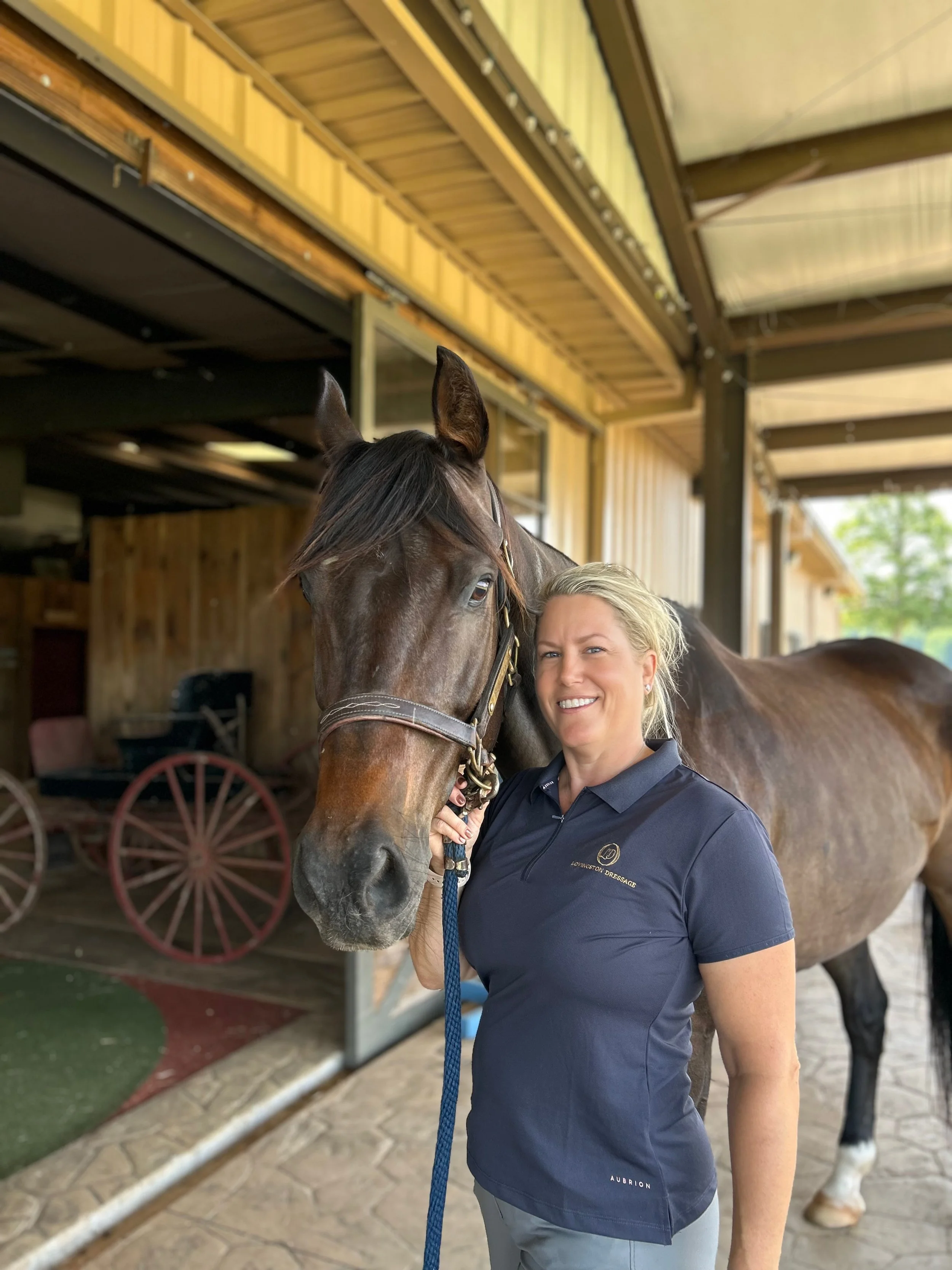 A woman in a navy polo shirt standing next to a brown horse indoors, with a wooden barn ceiling and a red wagon in the background.