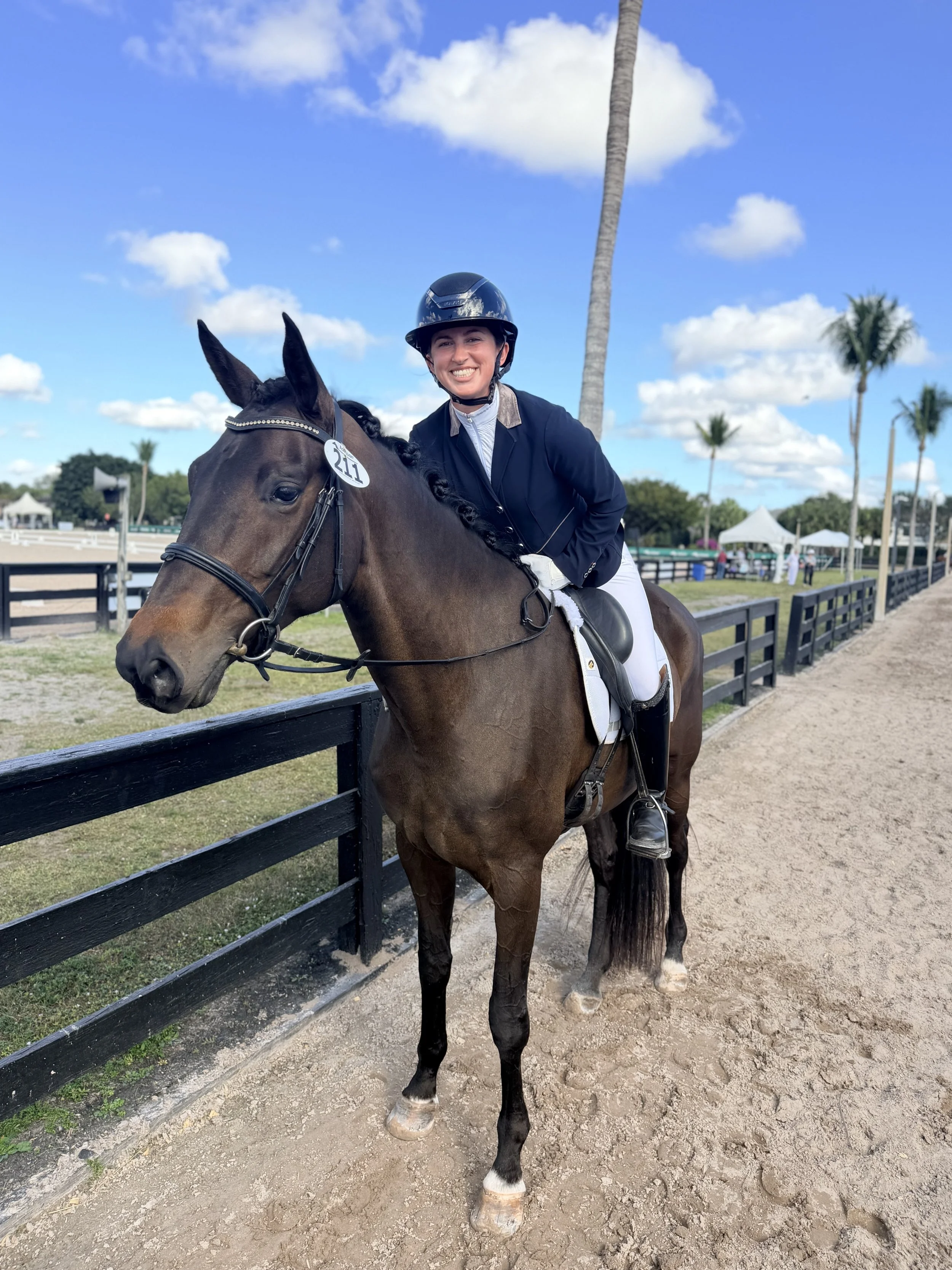 A woman in equestrian attire riding a brown horse on a dirt path at a horse racing or riding event, with palm trees and blue sky with clouds in the background.