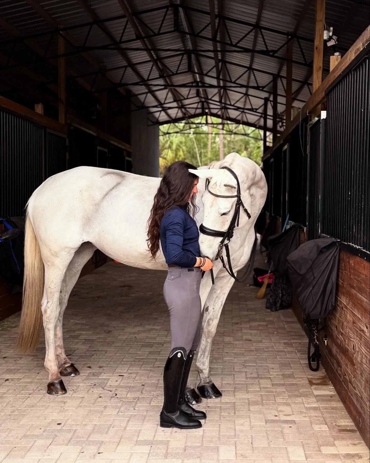 A woman standing with her head against a white horse inside a stable. The woman is wearing riding boots, gray breeches, and a navy blue shirt, and the horse is wearing a bridle. The stable has wooden walls and black barred stalls, with a brick floor 