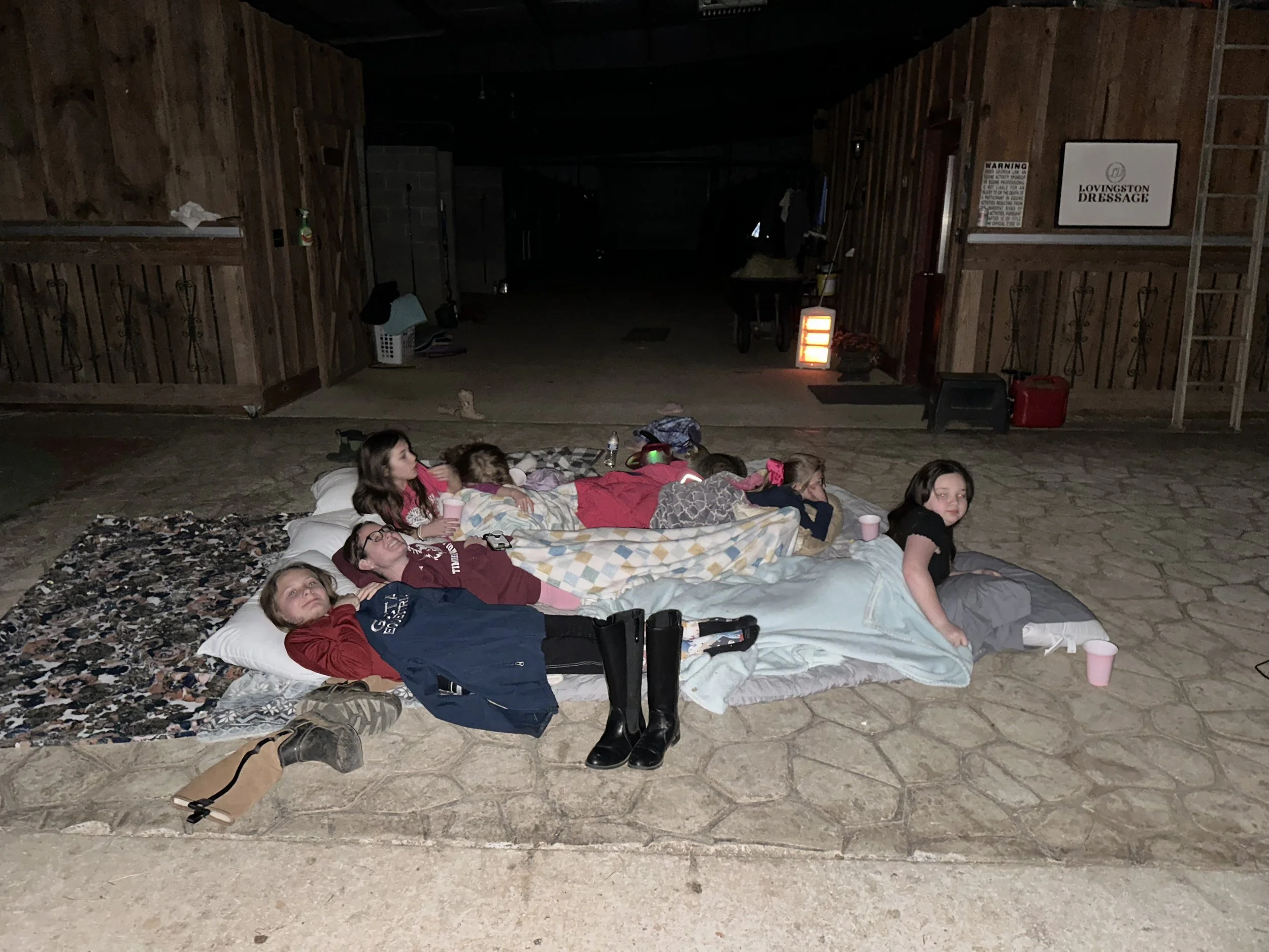 Group of children lying on mattresses and blankets on a concrete floor inside a wooden barn or shed, some holding cups, with a few boots and hanging coats nearby.