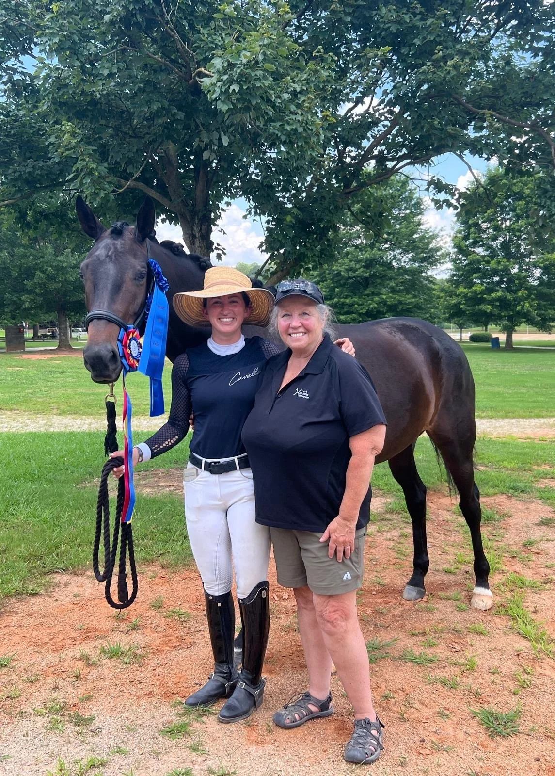 Two women and a dark brown racehorse under a large leafy tree. One woman is dressed in equestrian attire with a wide-brimmed straw hat, holding the horse's reins and wearing a blue ribbon. The other woman wears a black polo shirt and shorts. They are