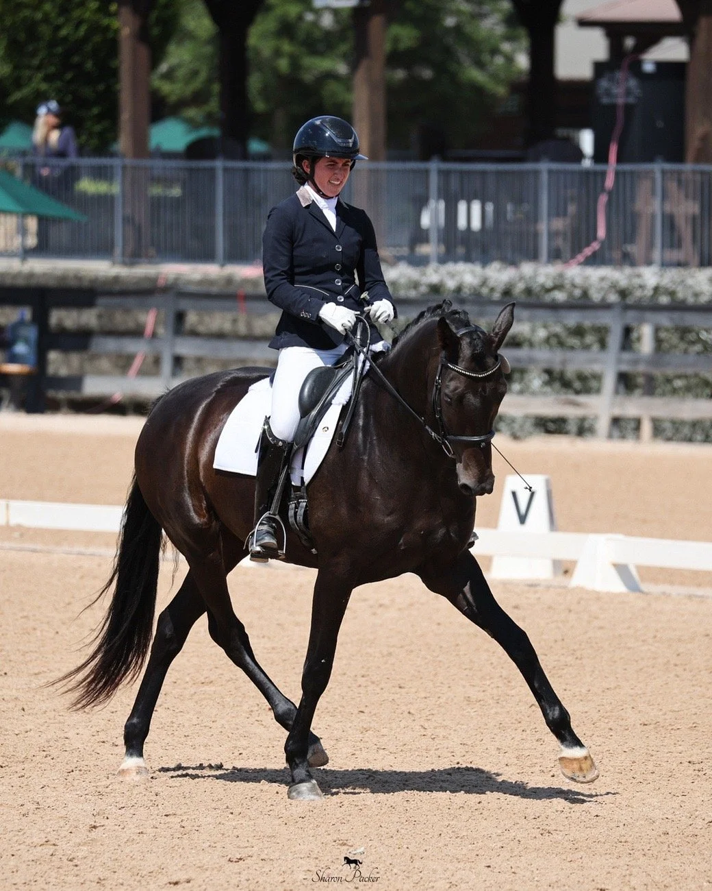 A woman riding a dark brown horse in a dressage arena during a competition, wearing a black helmet, navy blue riding jacket, white breeches, and gloves.