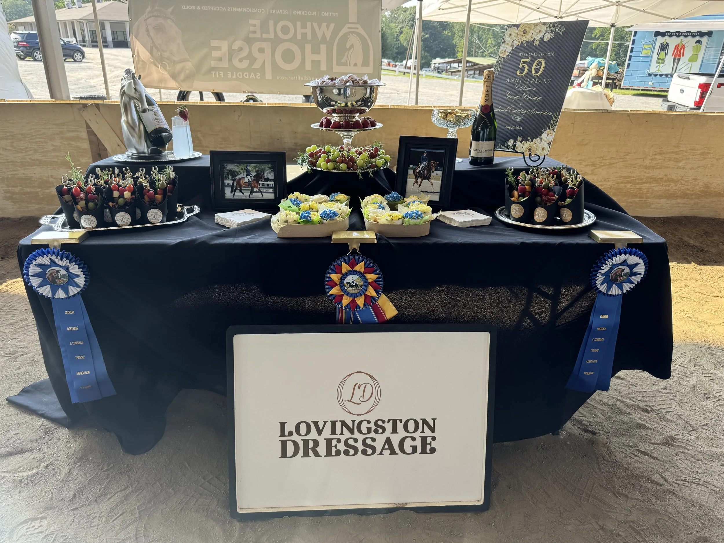 Display table at an event with awards, framed photographs, cupcakes, grapes, a bottle of Moët & Chandon champagne, and decorative ribbons, celebrating Lovingston Dressage's 50th anniversary.
