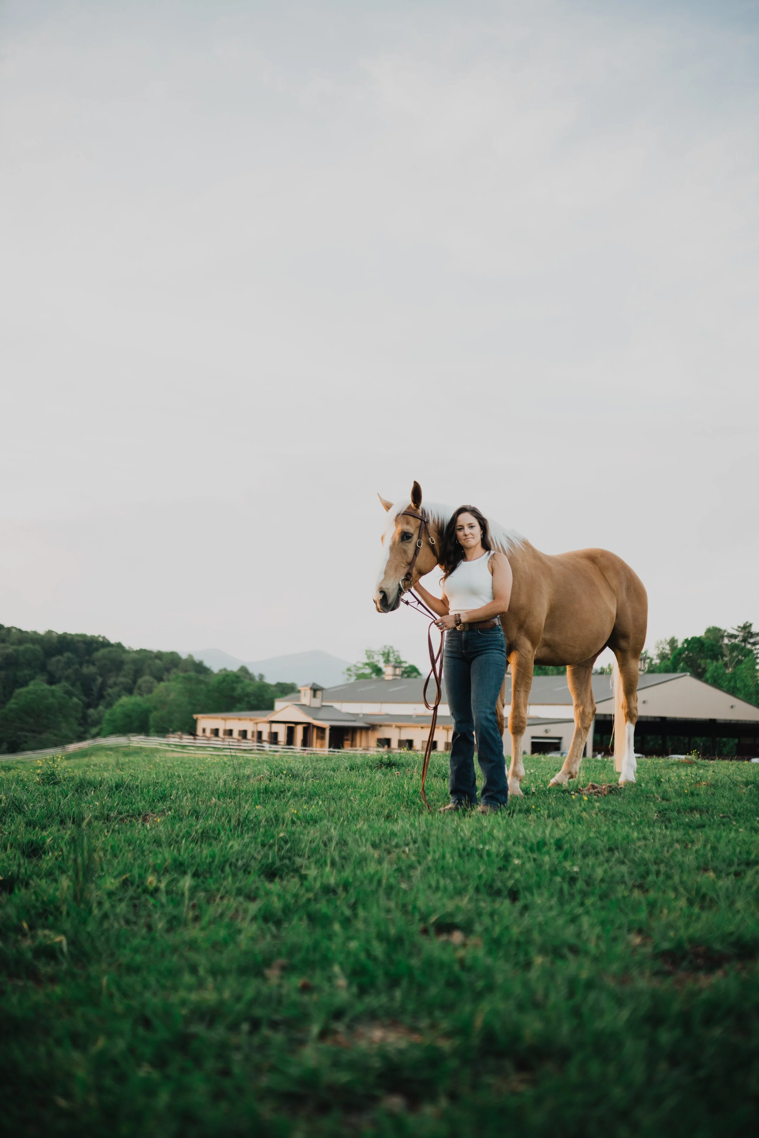 A woman standing in a grassy field holding a horse by the reins. There is a large building and hills in the background, under a partly cloudy sky.