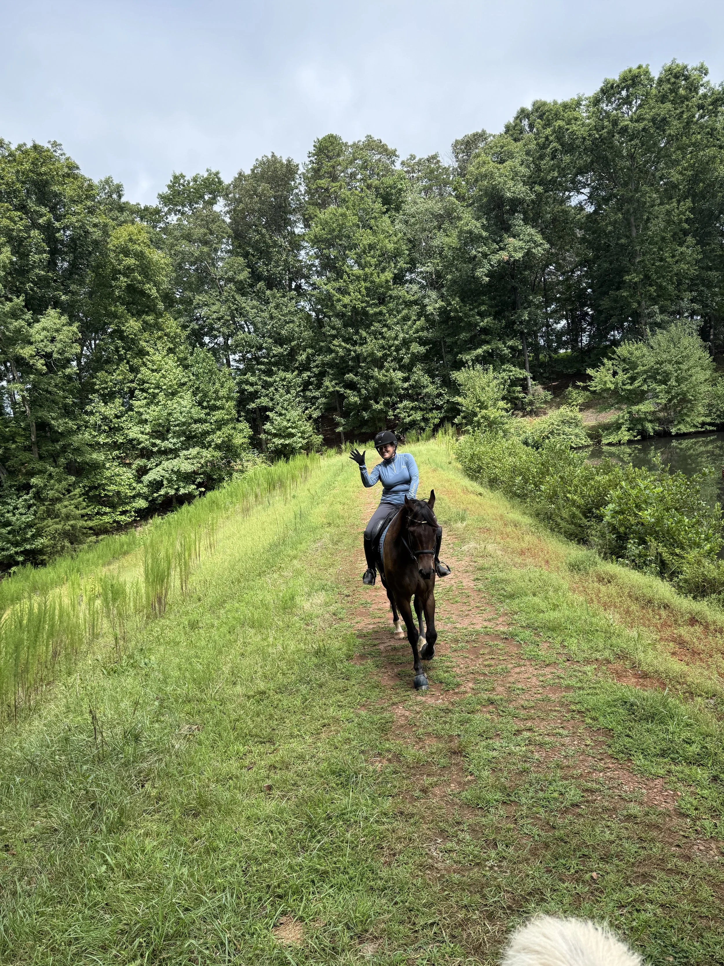 A woman riding a horse on a grassy trail surrounded by green trees, waving at the camera, with a lake to the right and a clear sky above.